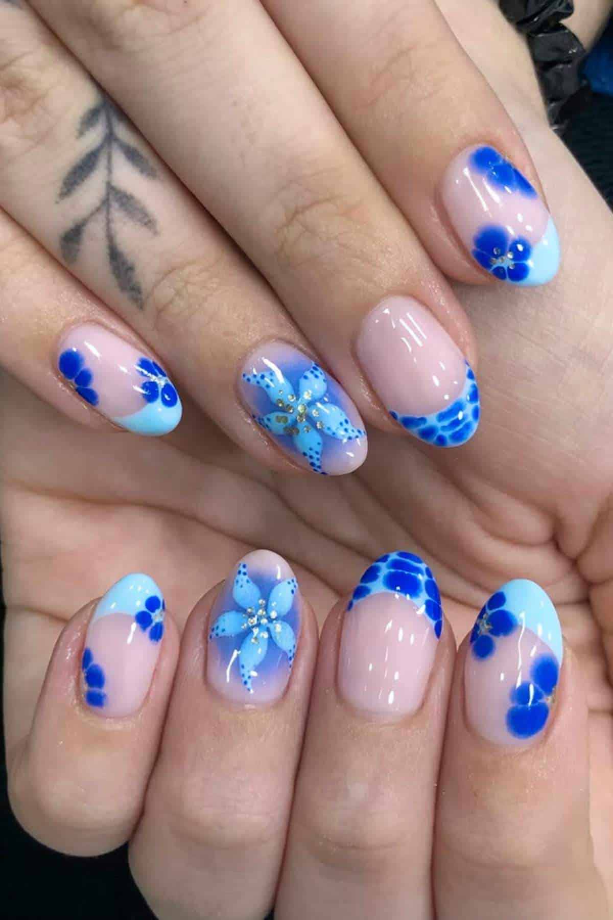 Close-up of hands with almond-shaped nails featuring light blue French tips and blue floral nail art designs on a natural pink base.