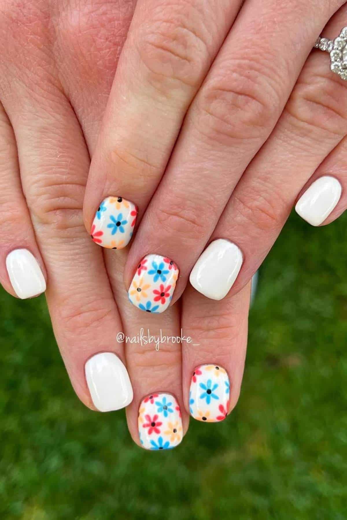 Hands with short, square-shaped nails; most nails are painted white, while a few feature a multicolored floral design on a light background.