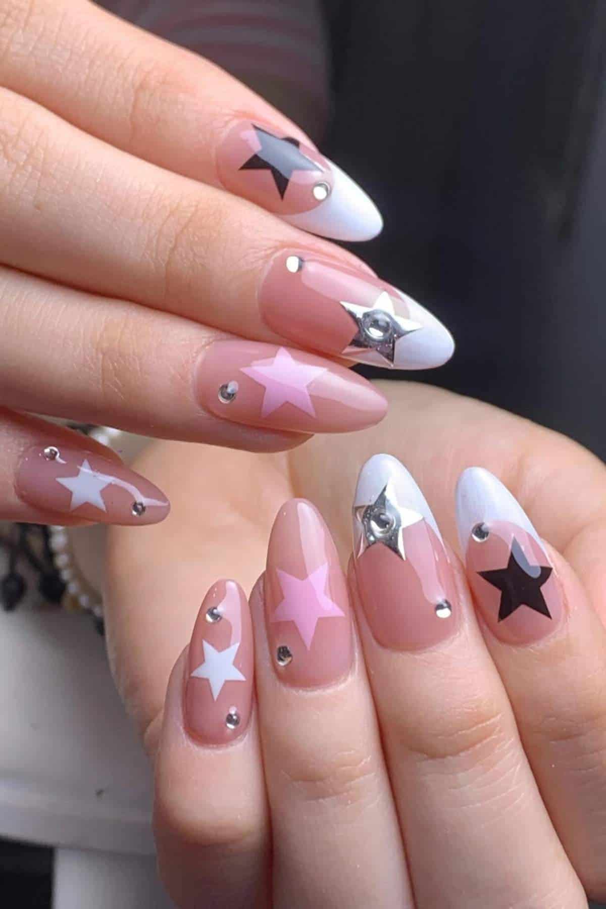 Close-up of hands with almond-shaped nails featuring pink, white, and black star designs, silver studs, and white tips on a nude base.