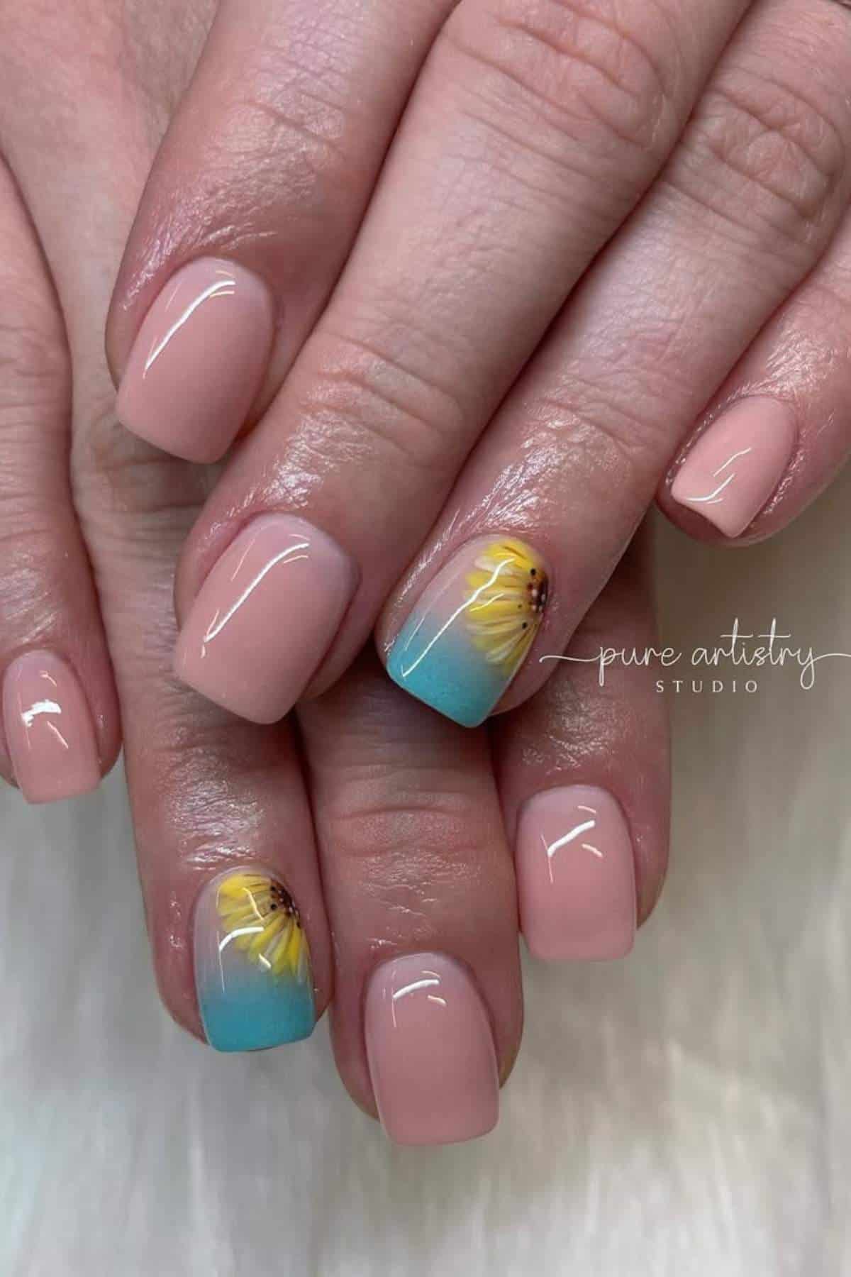 Close-up of manicured hands with light pink nails; two accent nails feature a sunflower design on a blue gradient background.
