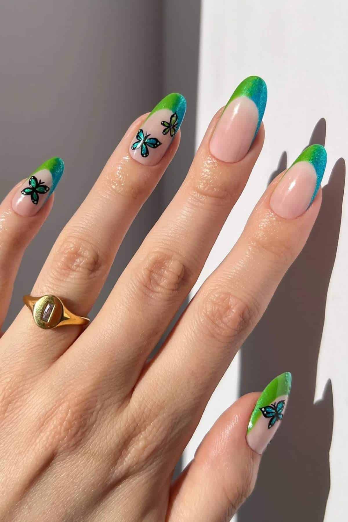 A hand with long nails featuring blue and green French tips and butterfly nail art, wearing a gold ring, shown in natural light.