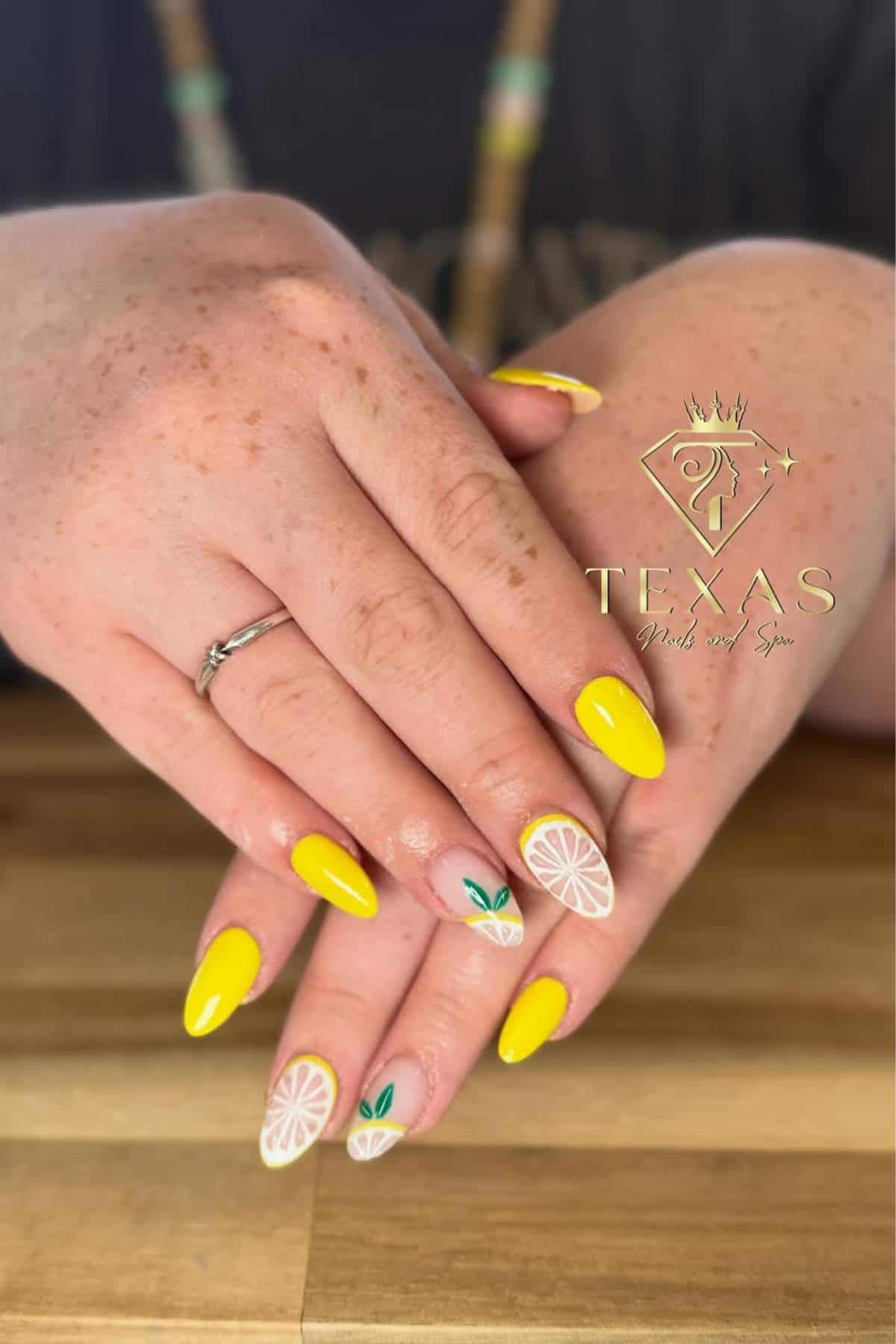 Hands with almond-shaped nails, painted bright yellow, some featuring lemon slice and leaf designs; Texas Nails and Spa logo in the corner.