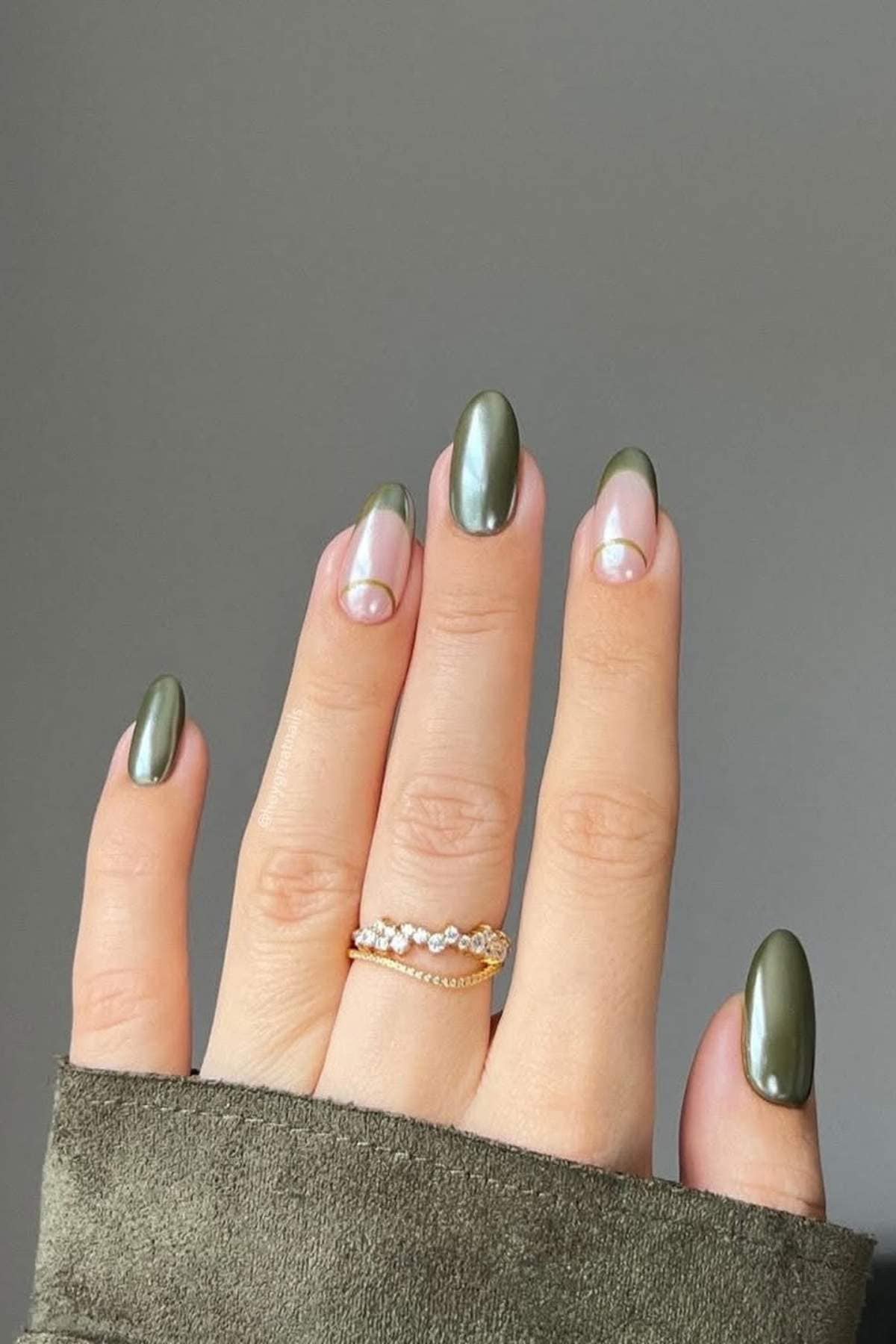A hand with almond-shaped nails; nails are painted metallic olive green with accent nails featuring a pearl and clear half-moon design. The person wears two gold rings.