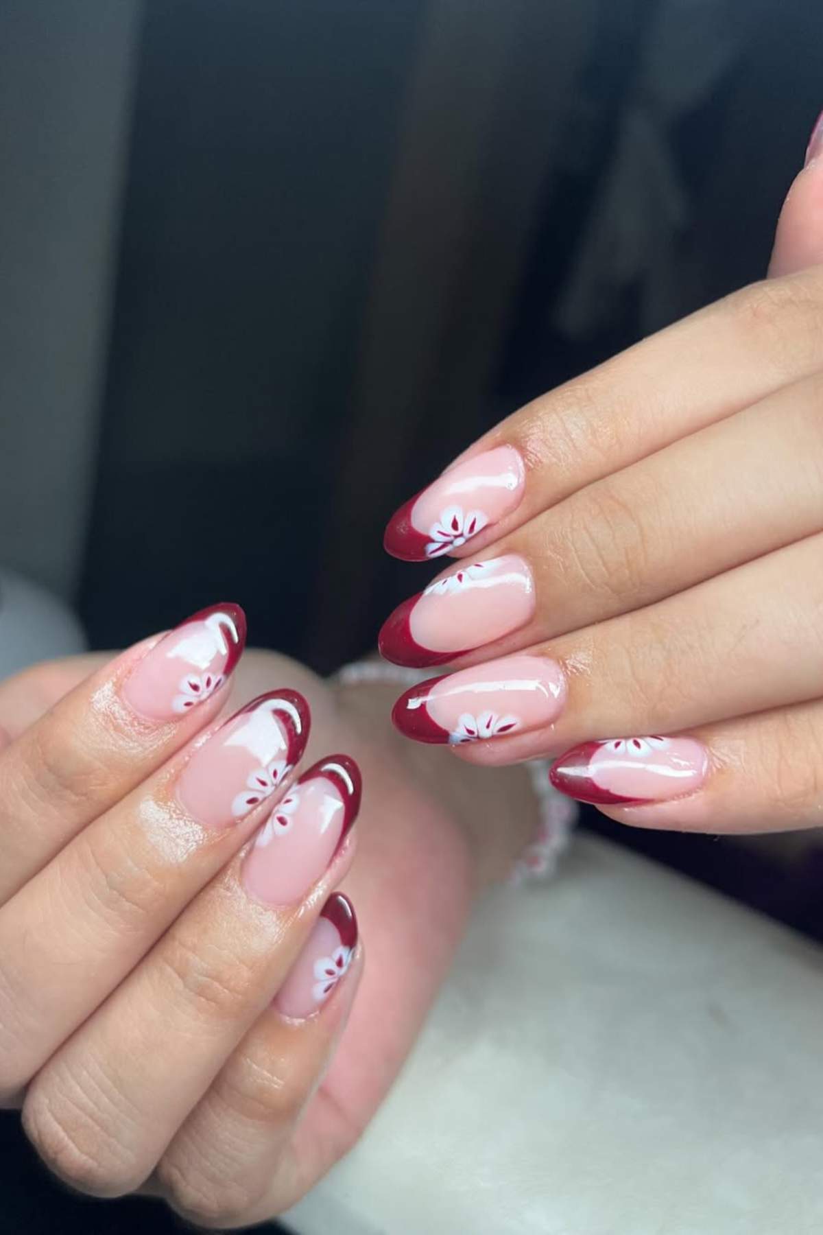 Hands with almond-shaped nails featuring red French tips and white flower designs on a nude pink base.