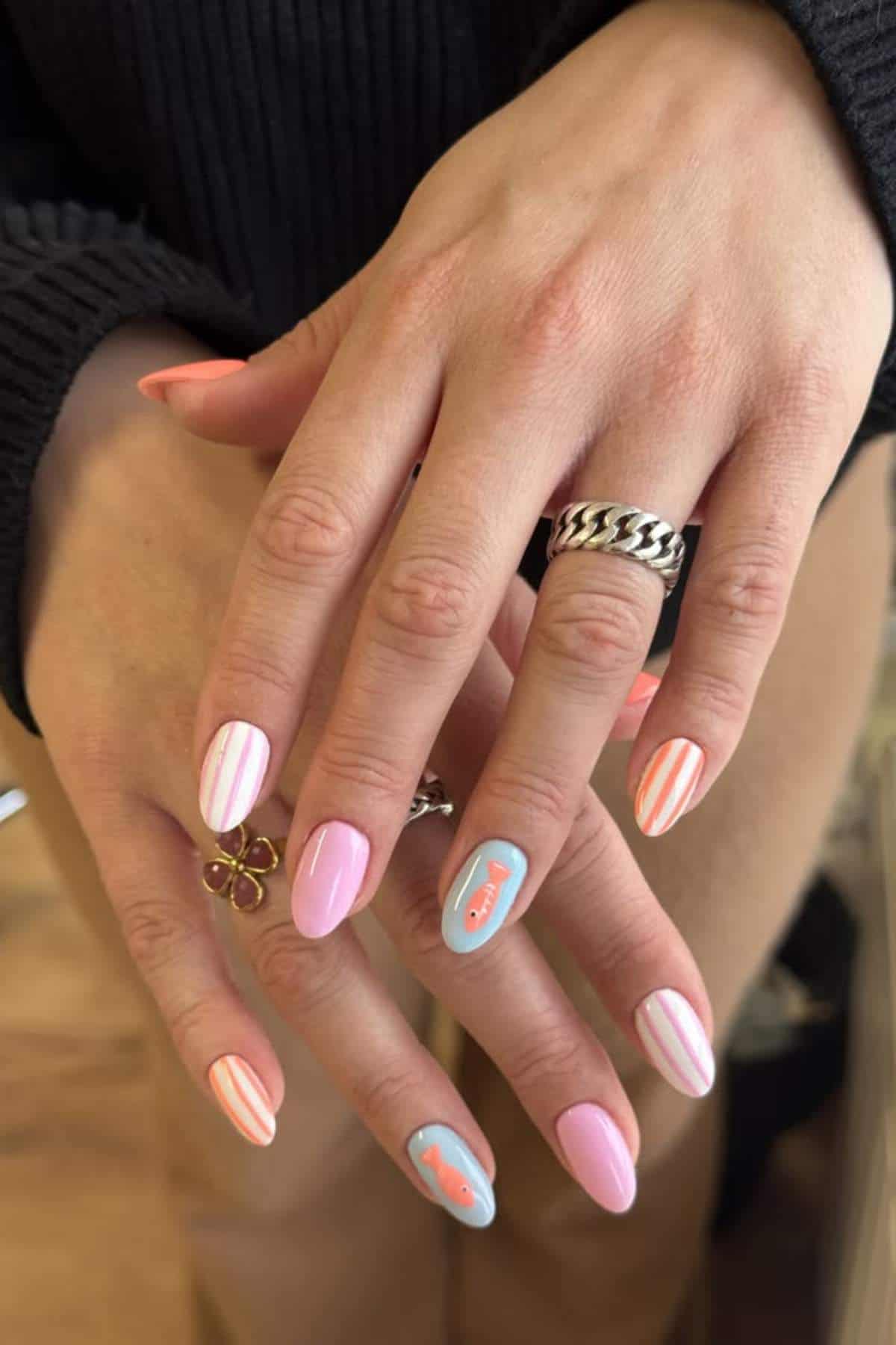 Hands with manicured nails featuring pink and white stripes, and blue accent nails with “love” and a heart design. Person is wearing rings and a dark long-sleeve top.