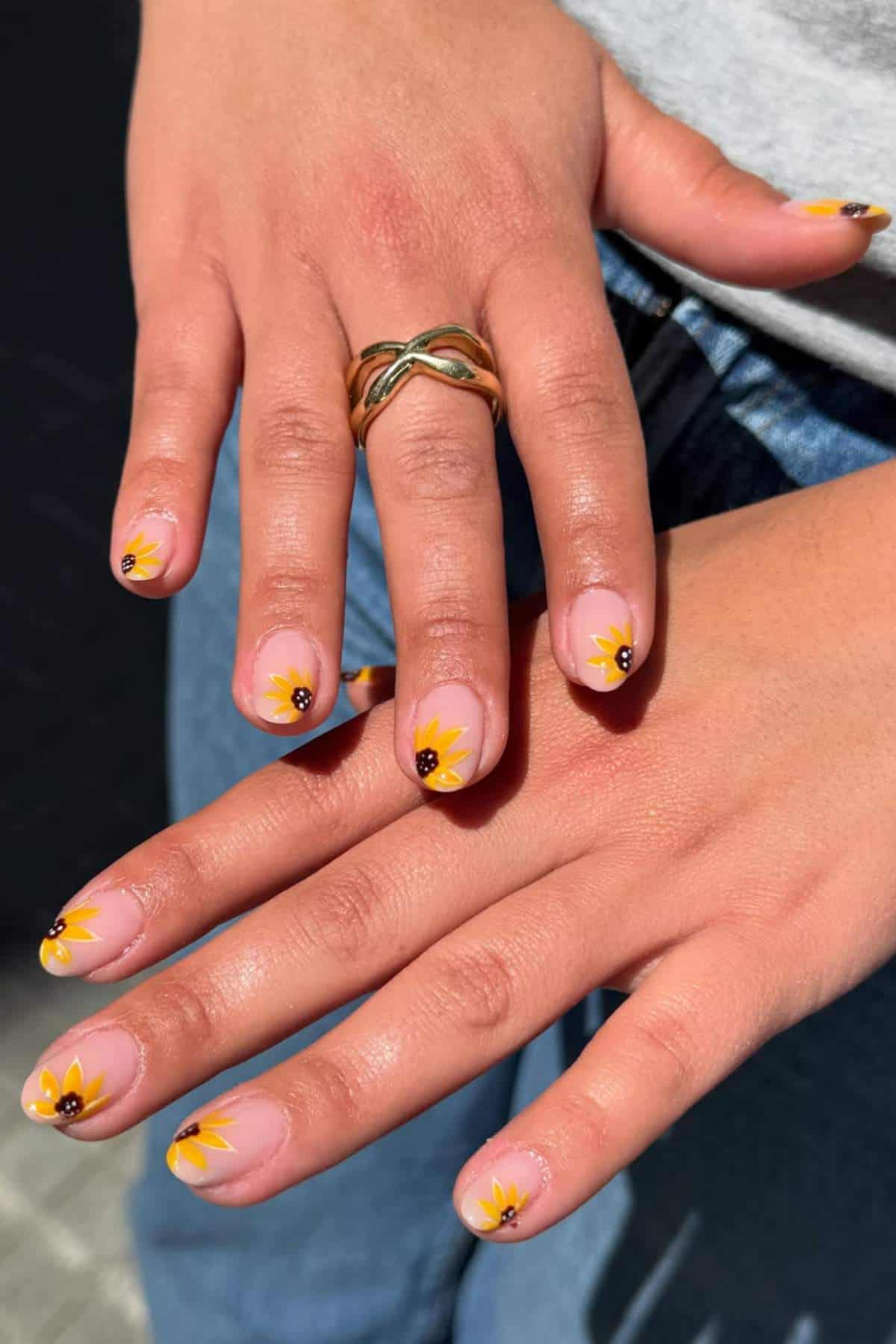 Close-up of hands with yellow sunflower nail art on short, natural nails. Person is wearing a gold ring with a twist design on the index finger.