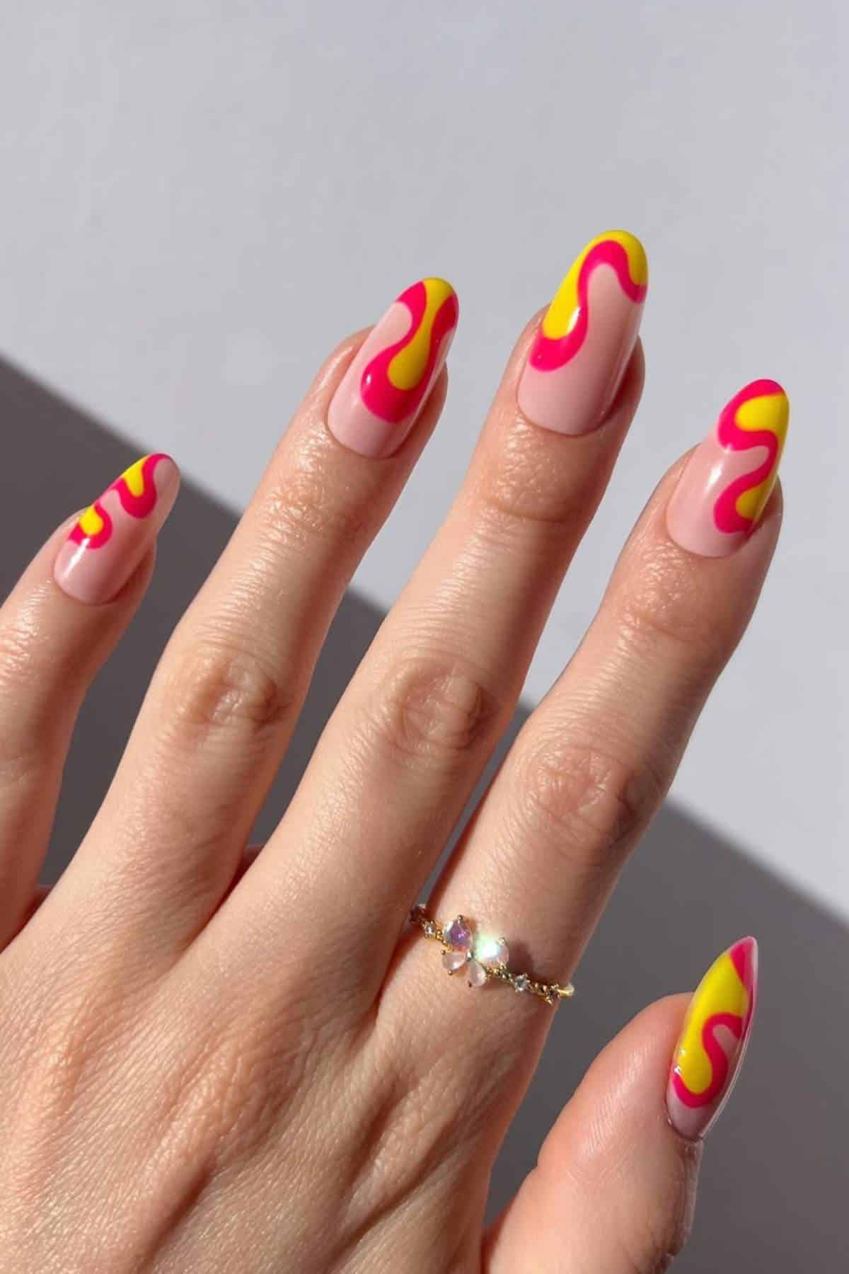 A hand with long almond-shaped nails featuring a pink base and yellow and red wavy designs, wearing a gold ring with small gemstones.