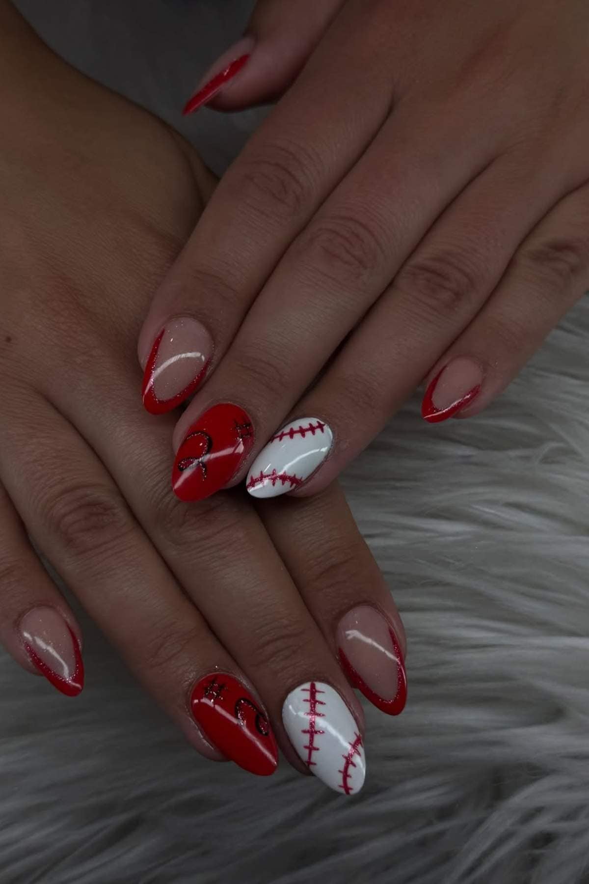 Hands with manicured nails featuring red tips, baseball stitching design, and glitter heart accents on a soft, textured surface.