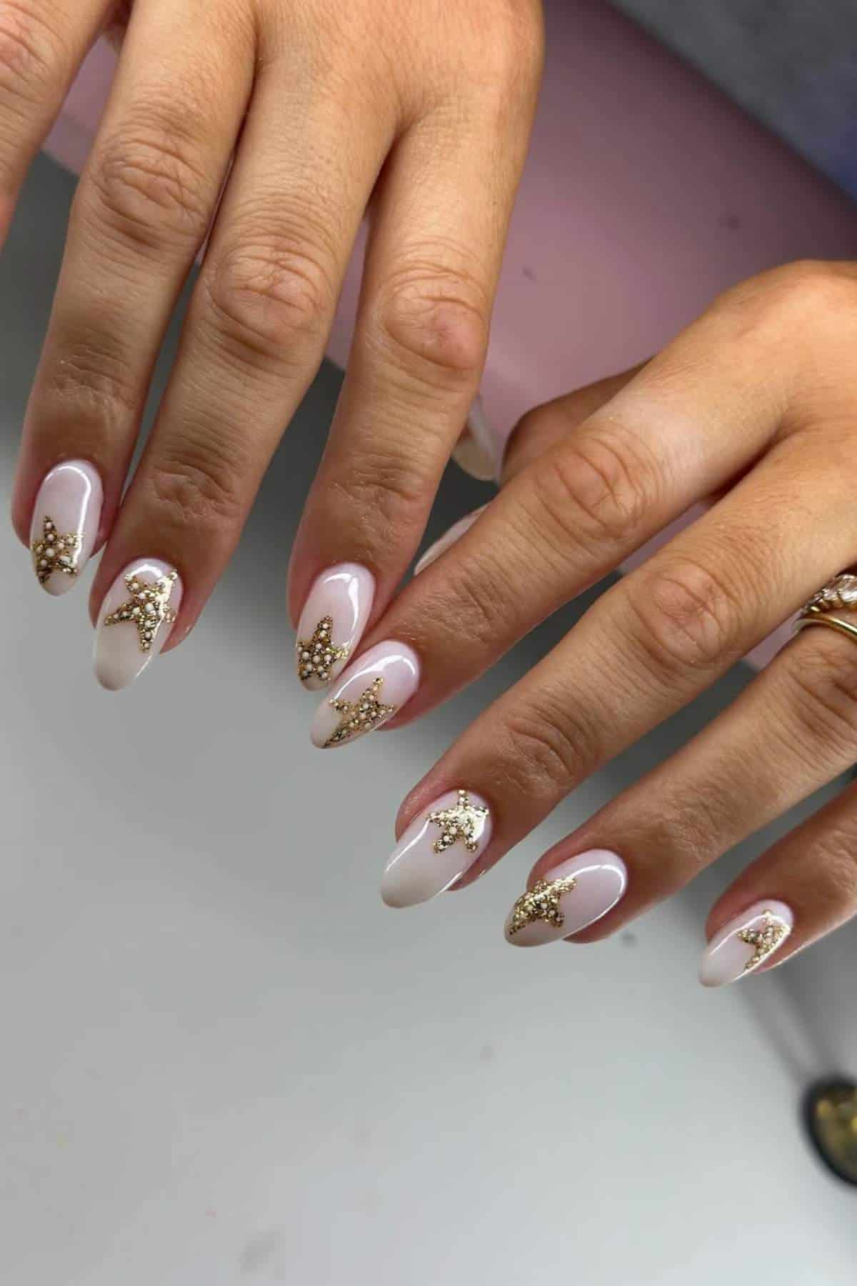 Hands with almond-shaped nails painted in a pale pink shade, featuring gold bead and sequin embellishments on each nail.