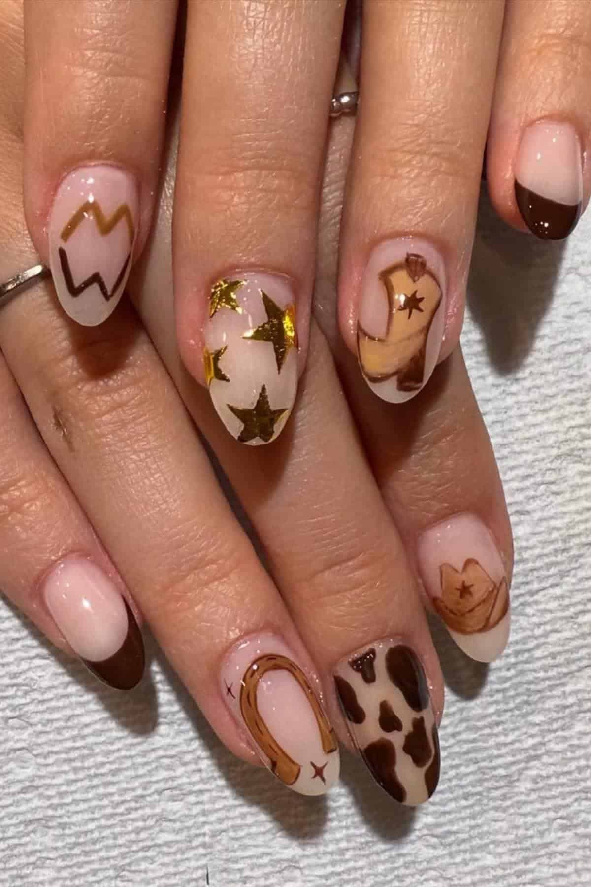 A hand with manicured nails featuring brown and gold Western-themed designs, including stars, cow print, and horseshoes on a nude base.