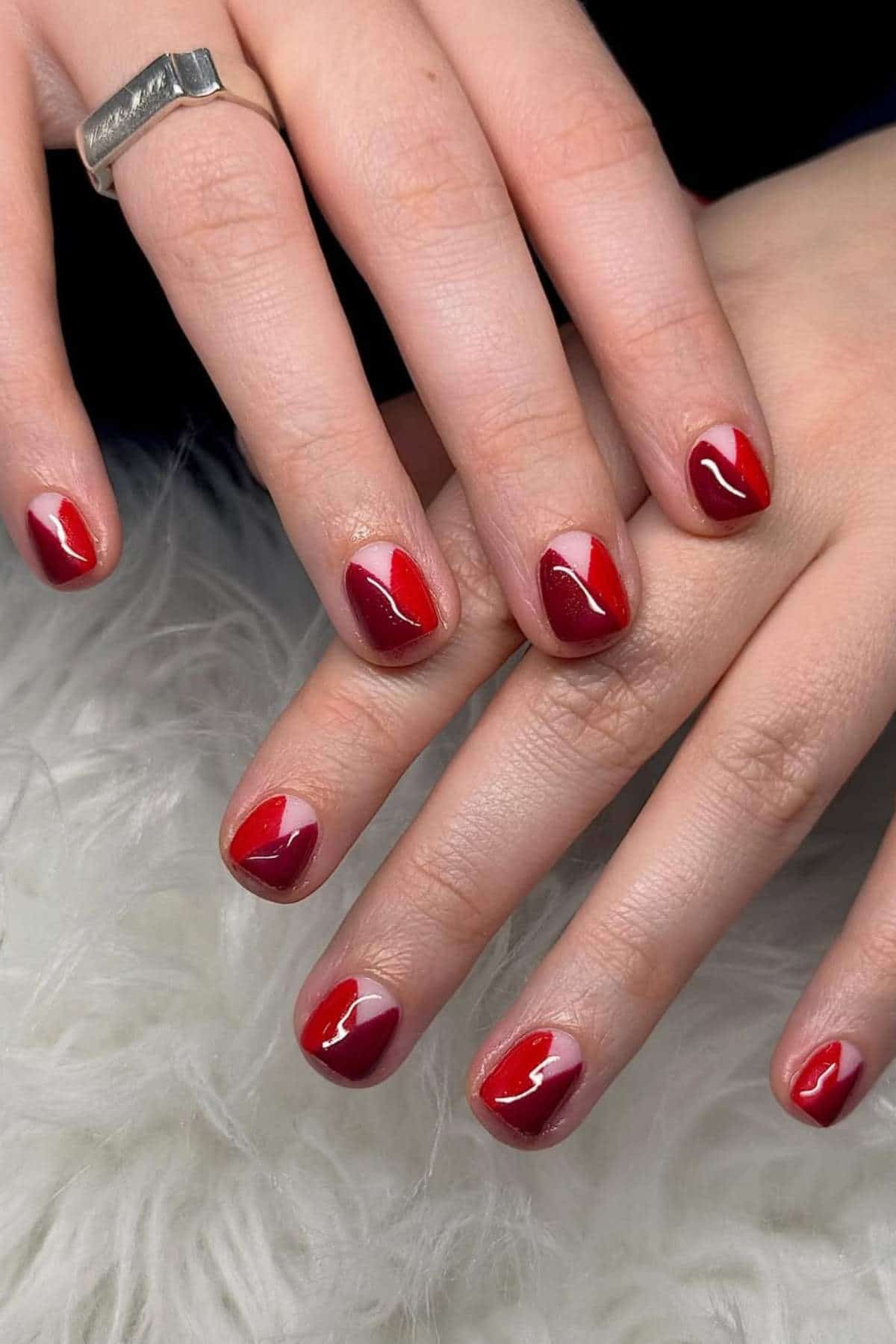 A close-up of hands with short, almond-shaped nails painted with red and pink heart designs on each nail. The person is wearing a silver ring.