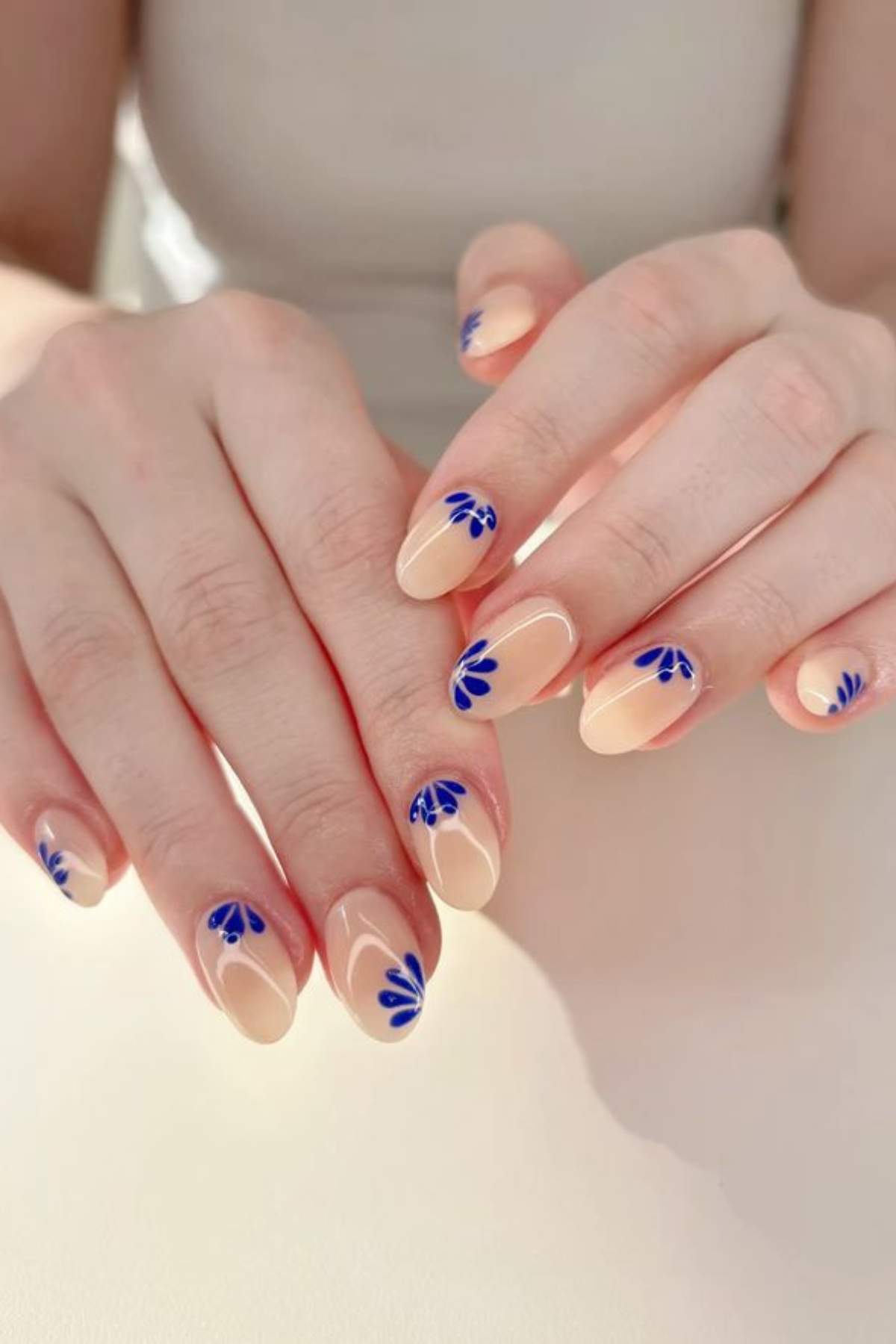 Hands with almond-shaped nails painted in a nude color, featuring small blue abstract petal designs near the cuticles on each nail.