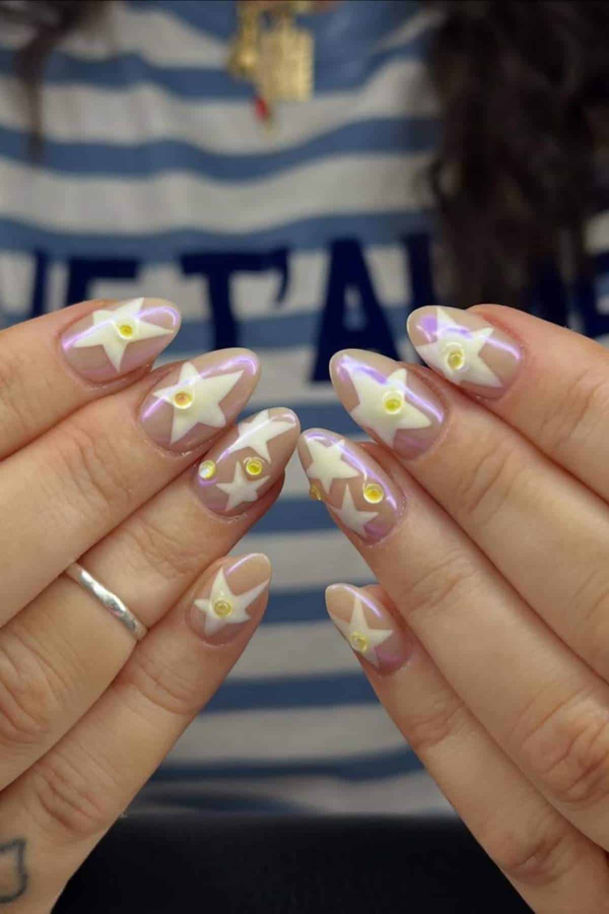 Hands with almond-shaped nails featuring white star designs, small yellow dots, and a glossy finish. Person is wearing a striped shirt and a silver ring on one finger.