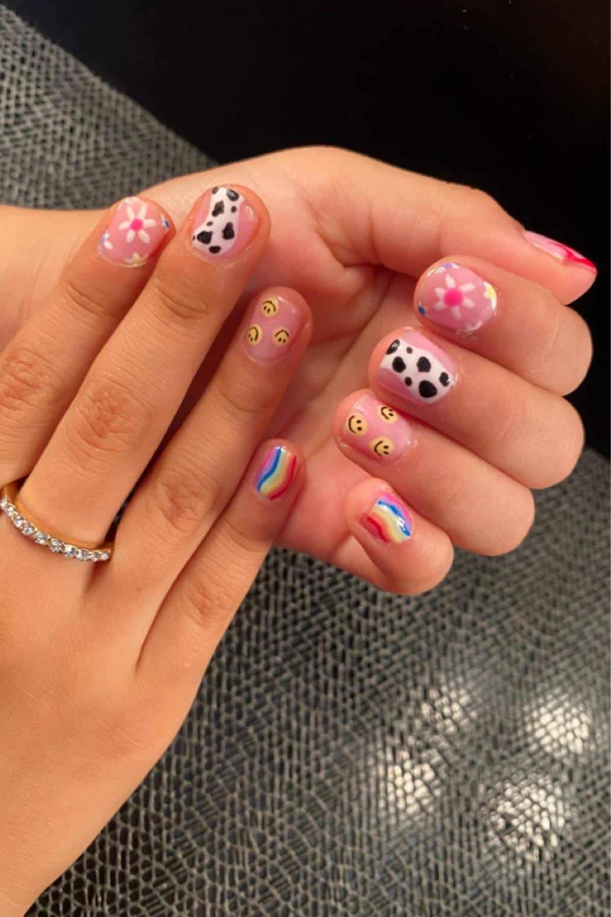 Hands with multicolored nail art featuring flowers, cow print, smiley faces, a rainbow, and wavy lines, shown against a textured gray surface.