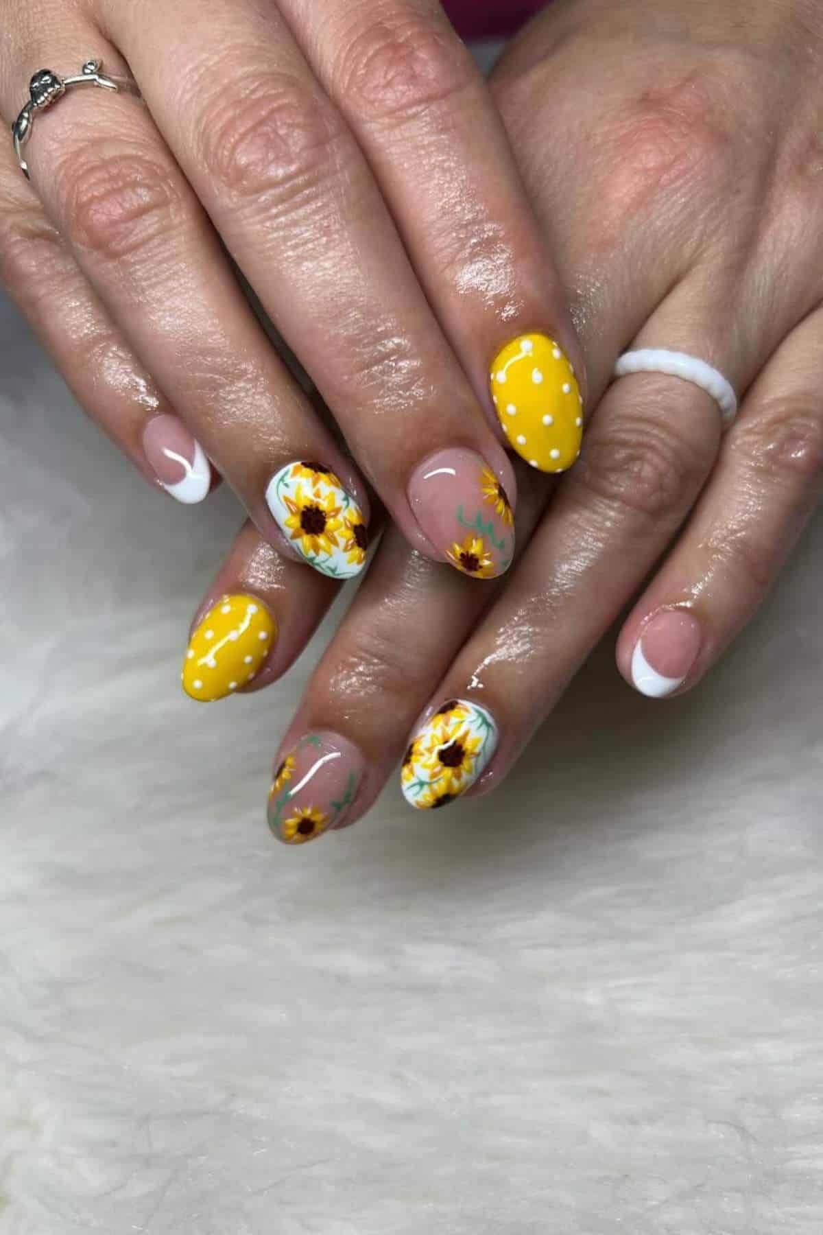 A set of manicured nails featuring yellow polish with white polka dots, white French tips, and sunflower designs on a light background.