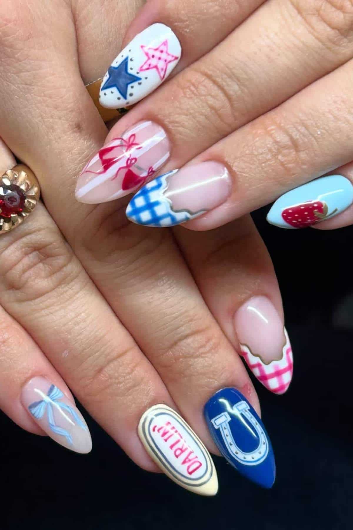 Close-up of hands with long, almond-shaped nails featuring colorful designs including stars, bows, check patterns, a horseshoe, a strawberry, and the word "darlin'.