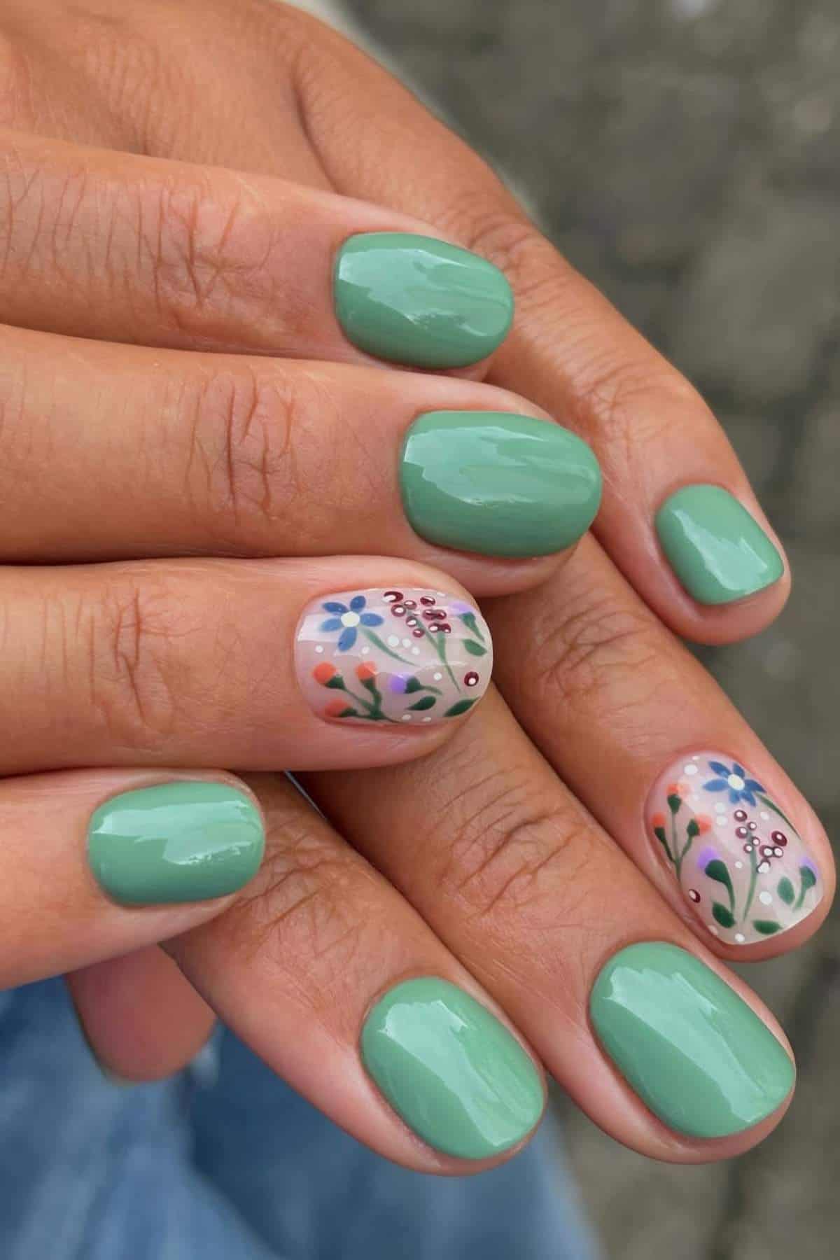 Close-up of hands with short, rounded nails painted green; two accent nails feature floral designs on a nude base.