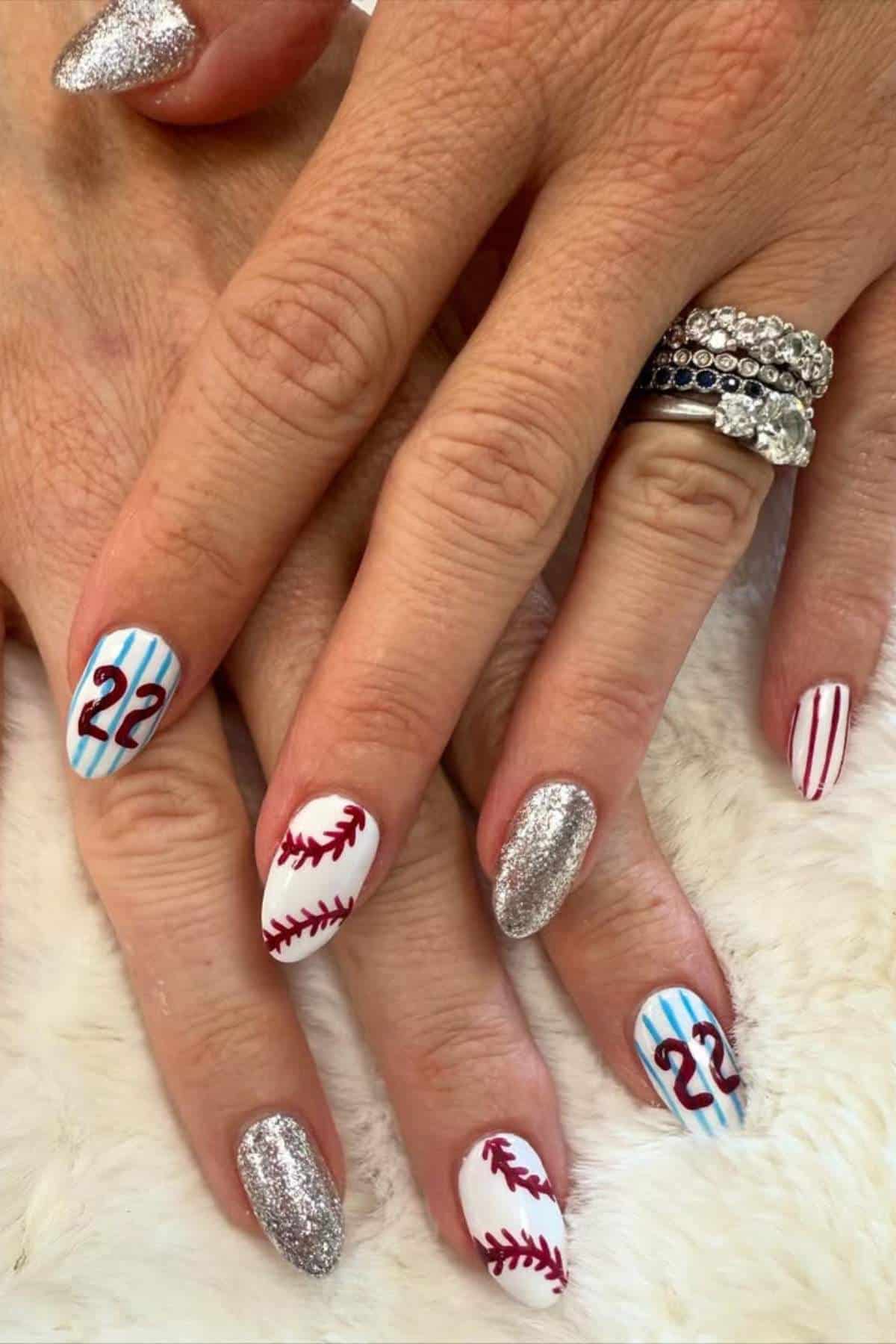 Hands with manicured nails featuring silver glitter polish, baseball stitching designs, blue pinstripes, the number 22, and red stripes. Rings are worn on the left hand.