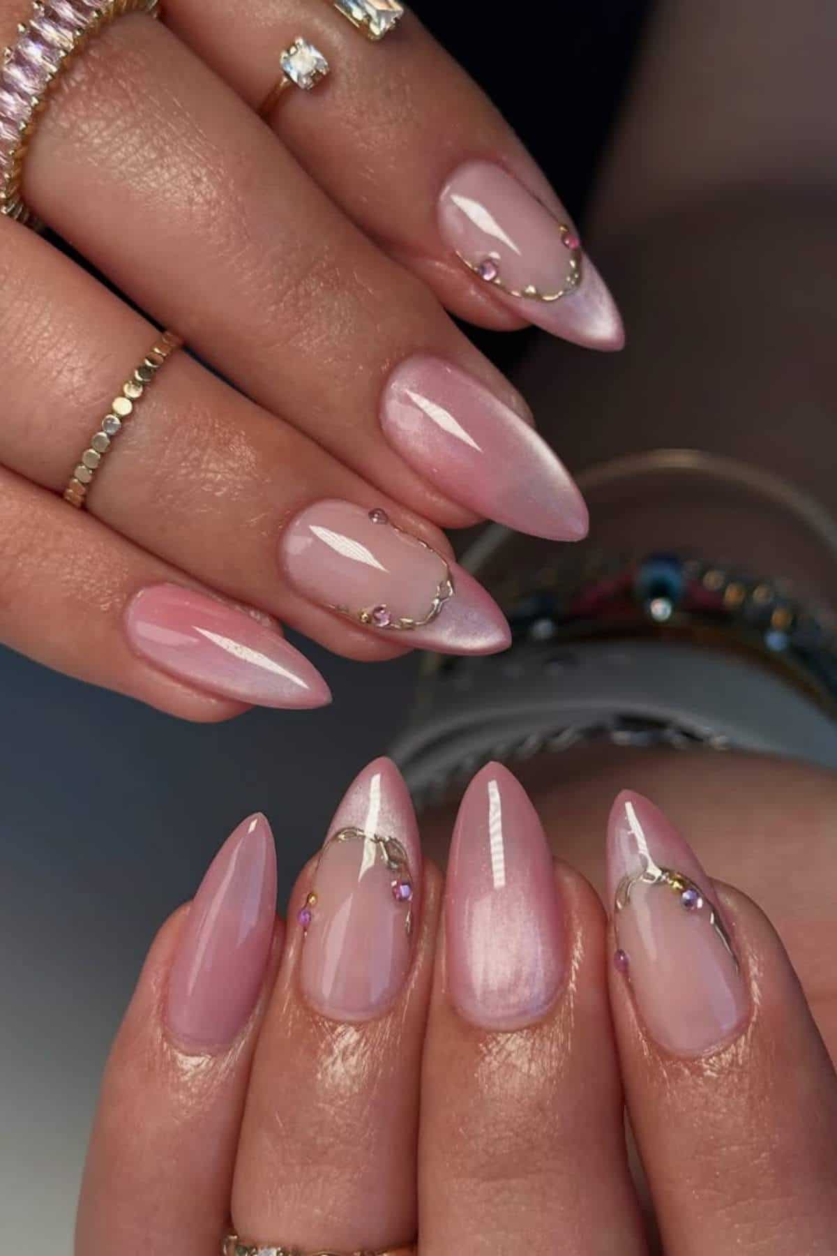 Hands with long, almond-shaped pink nails decorated with small gold chains and gemstones. Rings and bracelets are visible on the fingers and wrists.