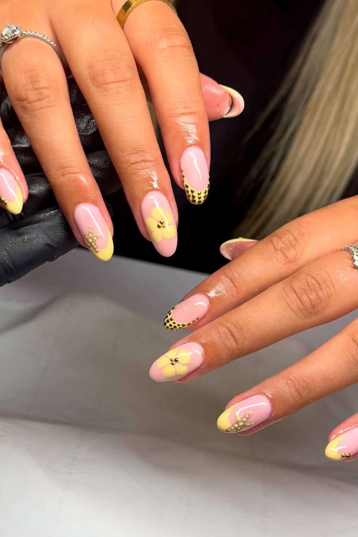 Hands with almond-shaped nails, painted in a pastel yellow and pink design with floral accents and black dotted tips on some fingers.