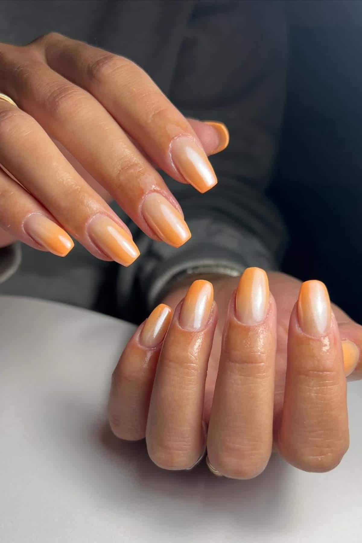 Close-up of hands with almond-shaped nails painted in a shiny gradient of white and orange shades.