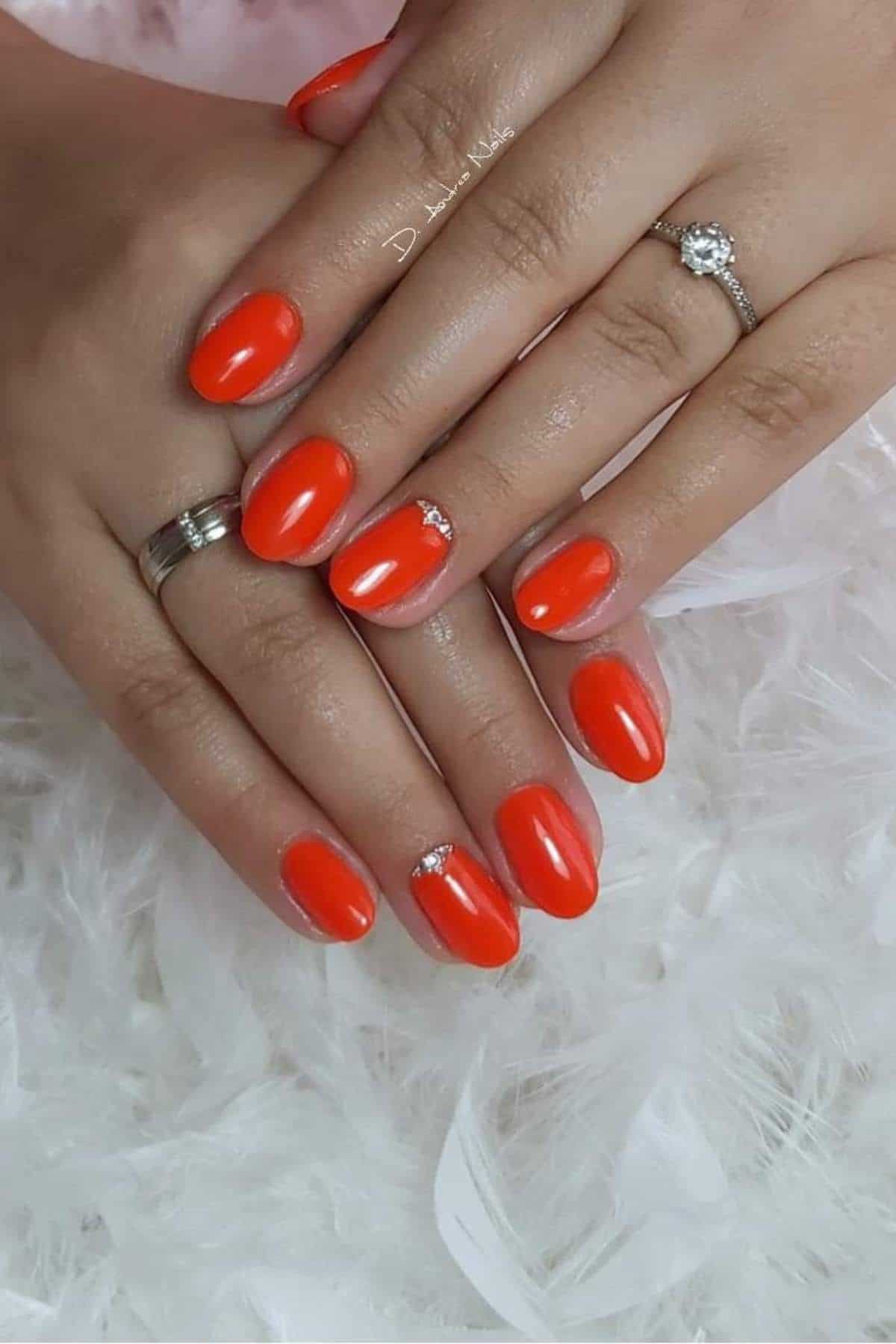 Hands with bright orange manicure, small silver accents on two nails, and rings on the ring fingers, displayed on a soft white textured surface.