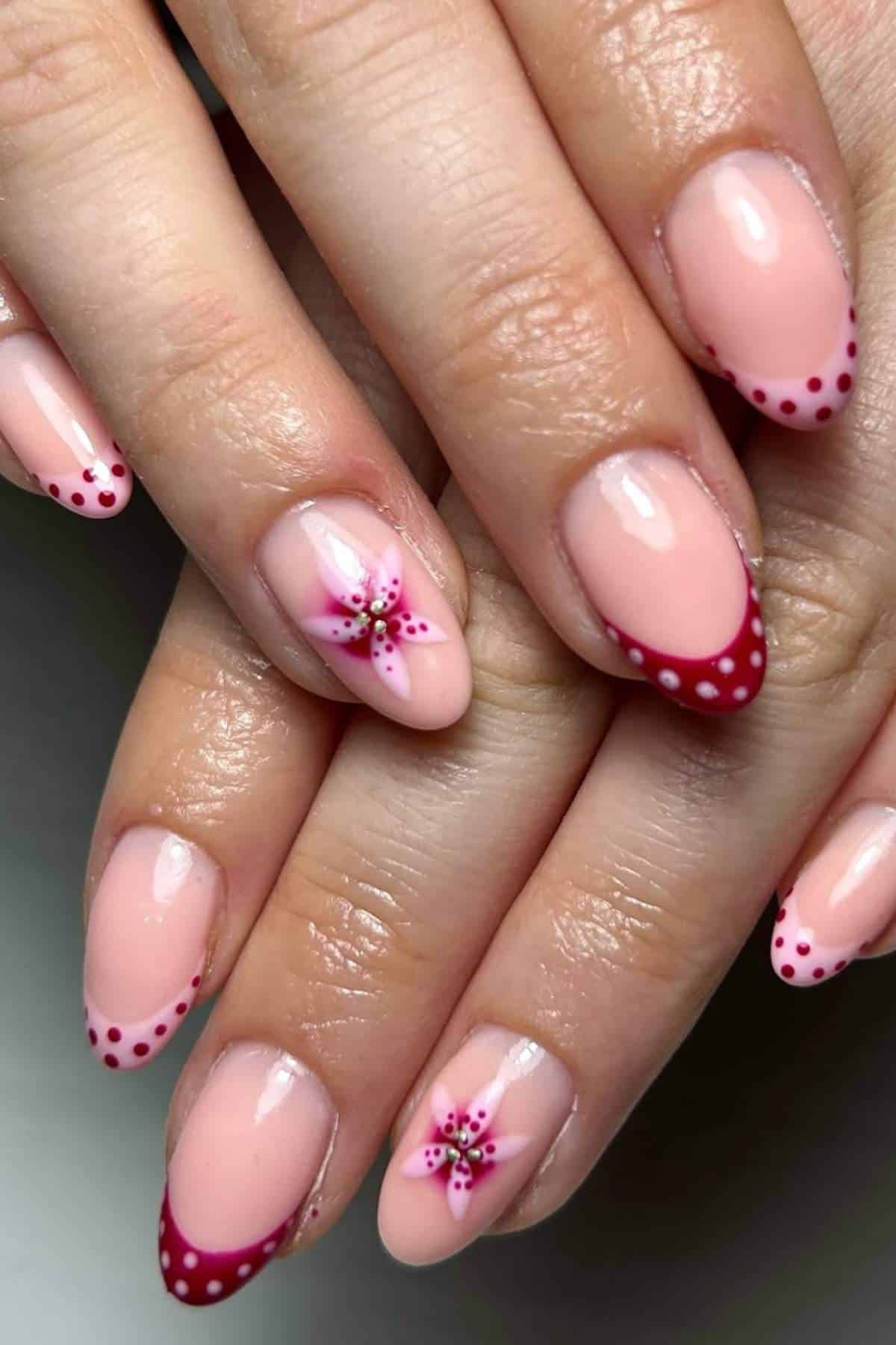 Close-up of hands with almond-shaped nails featuring nude polish, dark pink French tips with white polka dots, and pink flower nail art on two accent nails.