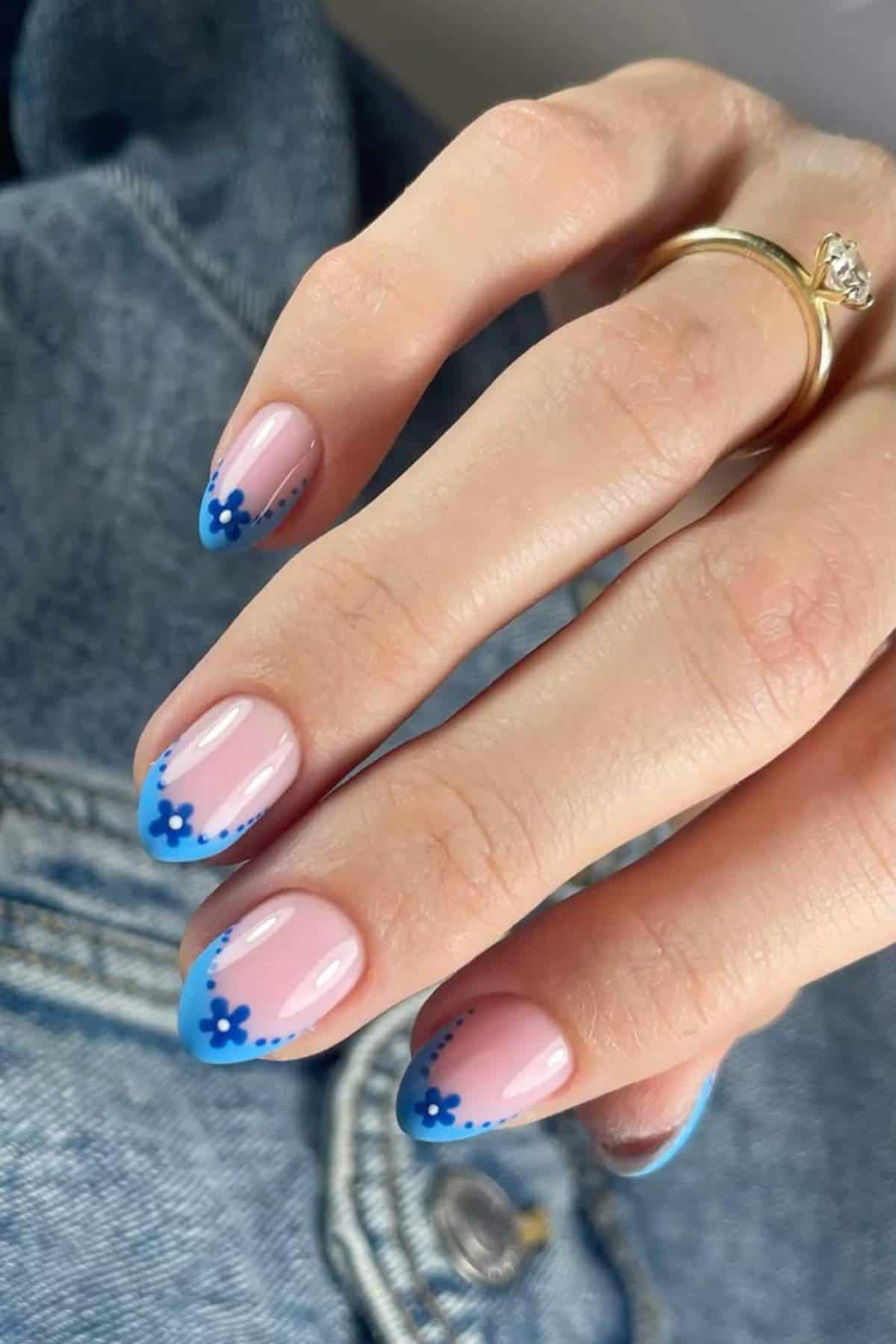 A hand with manicured nails featuring a nude base, blue floral designs, and blue French tips, shown against a denim jacket background.