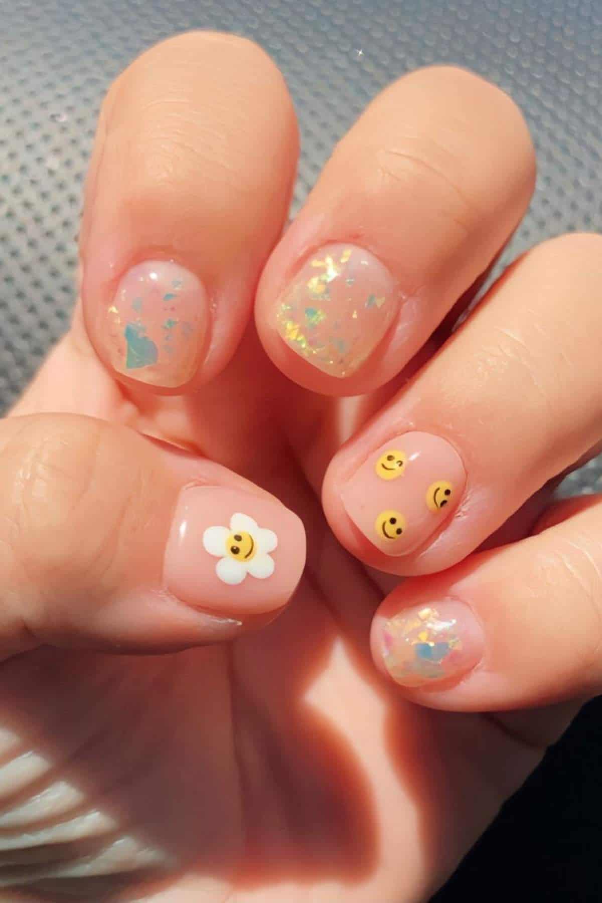 A hand with short, rounded nails features clear polish with iridescent flakes, smiley face decals, and a white flower with a smiley face design on the thumb.