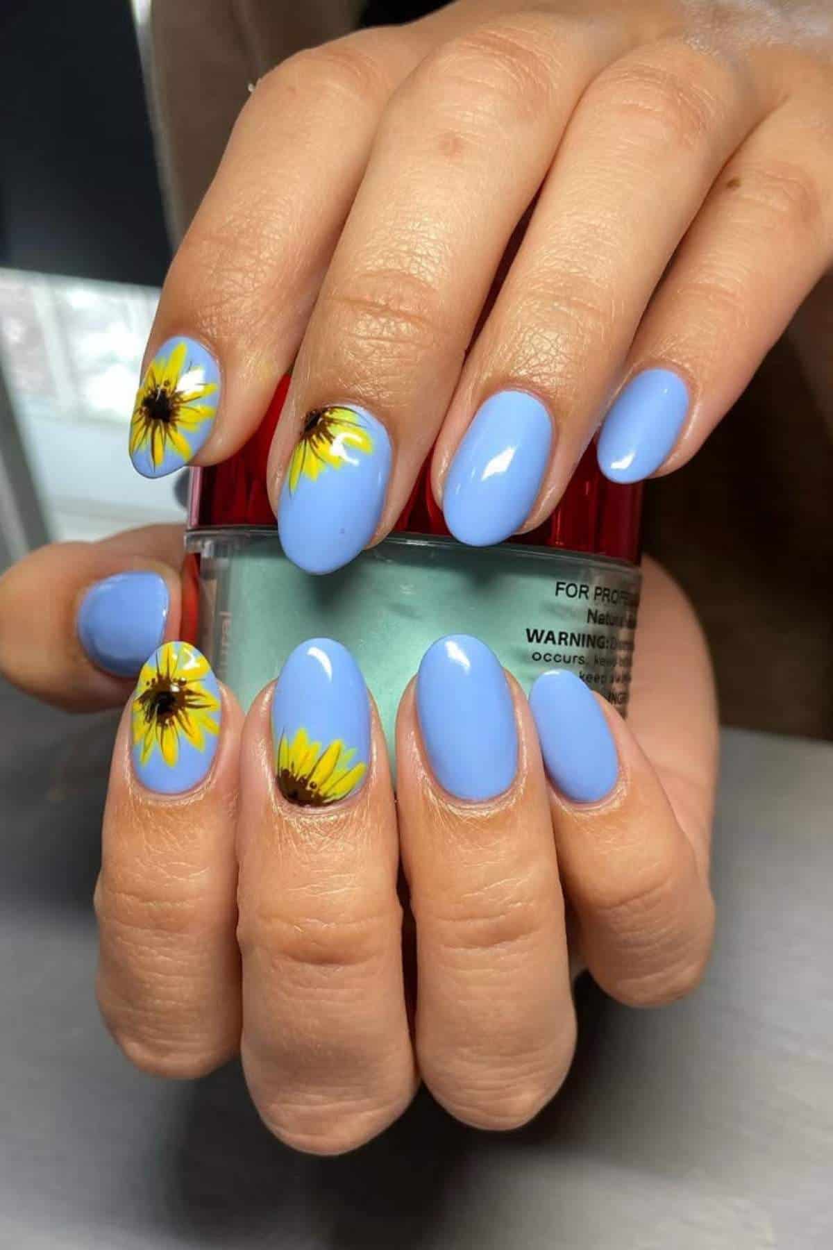 Hands with oval-shaped blue nails featuring yellow sunflower designs holding a small jar with a red lid.