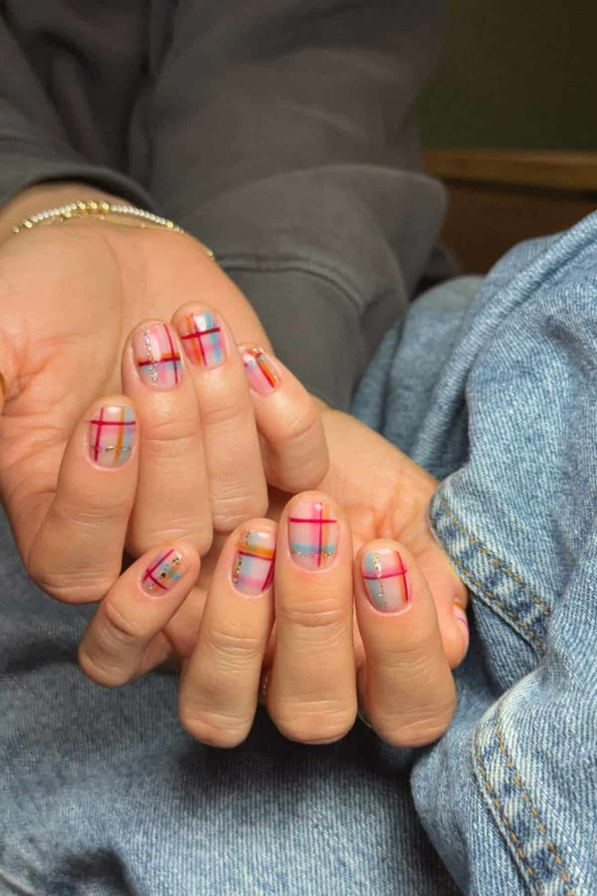Close-up of hands with short nails featuring colorful plaid and dot nail art on a nude base. Hands rest on denim fabric and a dark sleeve is visible.