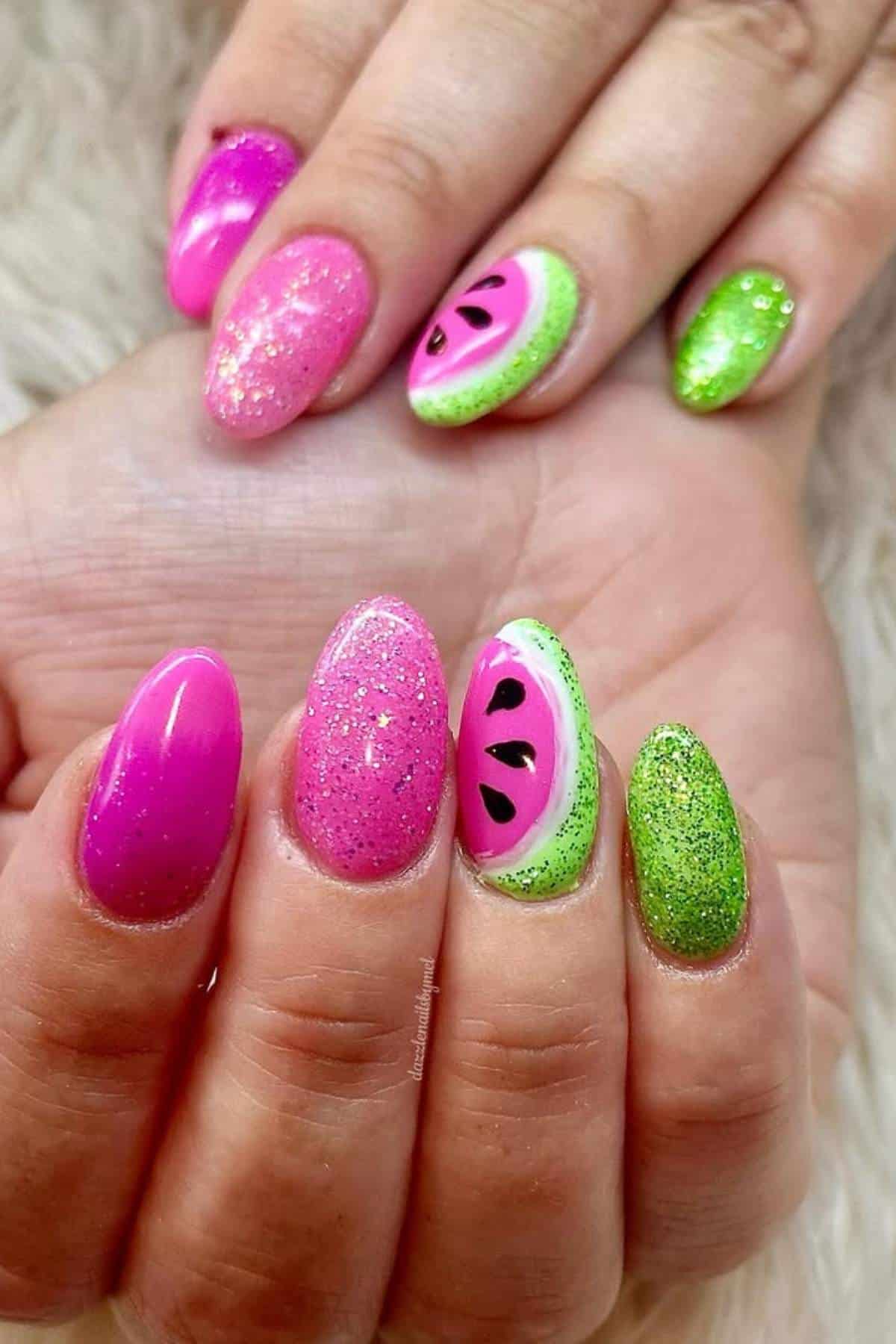 Hands with almond-shaped nails painted in pink and green, featuring glitter and a watermelon design on two accent nails.