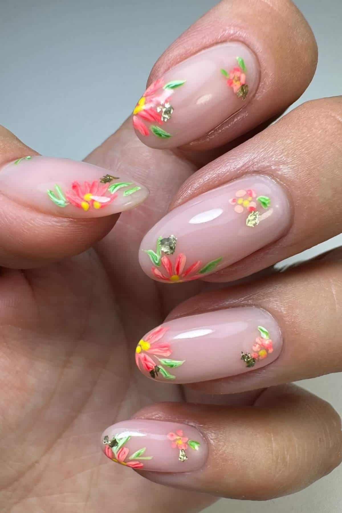 A hand with almond-shaped nails featuring a light pink base and decorated with small, colorful floral designs and touches of gold foil.