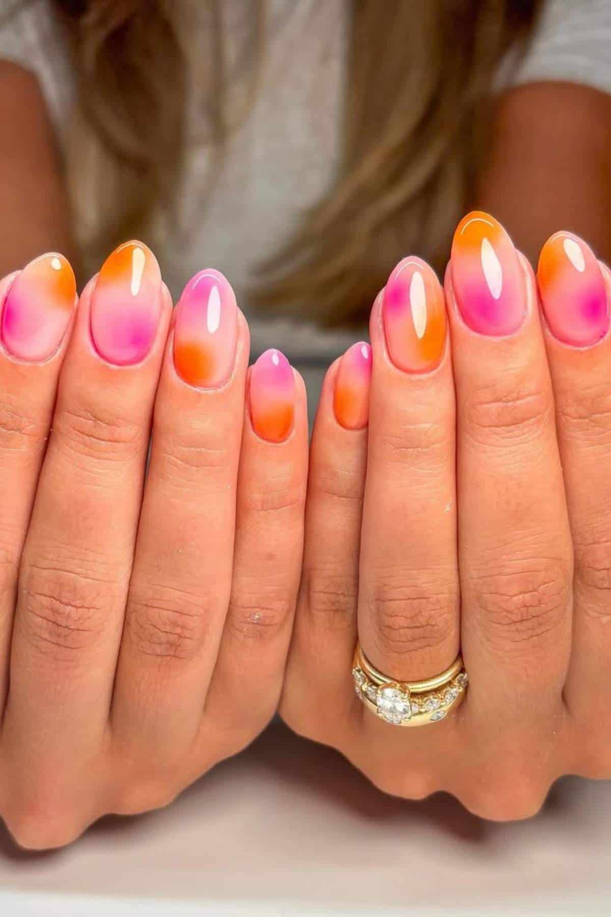 Close-up of hands with almond-shaped nails featuring a glossy pink and orange gradient design. The person wears gold rings, including one with a large gemstone.