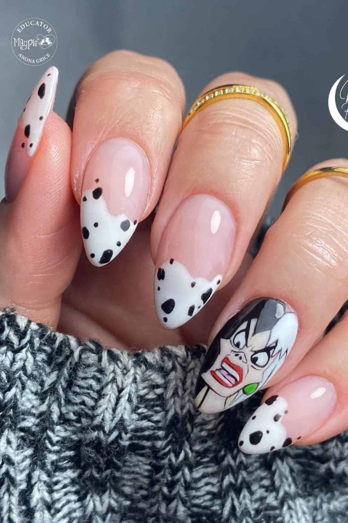 A hand with almond-shaped nails featuring Dalmatian print tips and an accent nail with a cartoon character face. The person is wearing gold rings and a gray knit sweater.