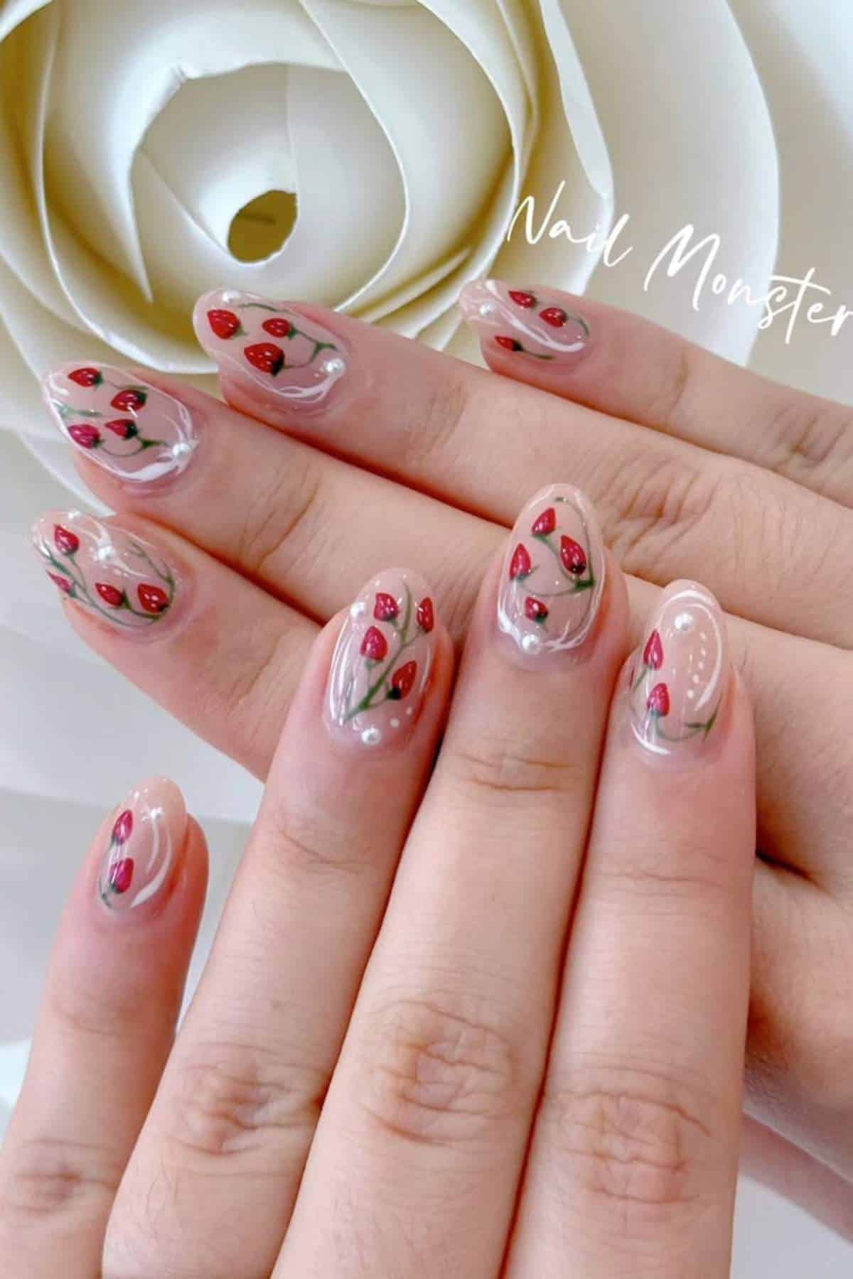 Close-up of hands with nude-colored nails featuring small red strawberry designs and green leaves, positioned in front of a large white paper flower.