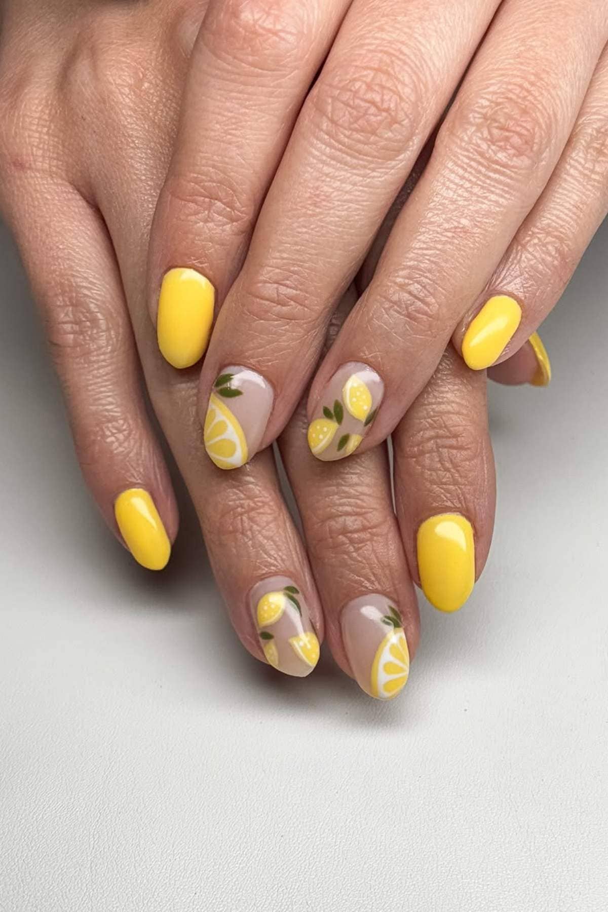 Hands with manicured nails painted yellow, some with lemon slice and leaf designs on a neutral base, resting on a white surface.