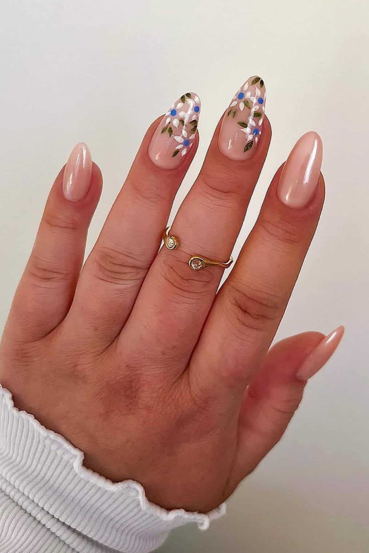 A hand with almond-shaped nude nails; two nails feature a floral design with blue and white flowers and green leaves. The person wears a gold ring with two small stones.
