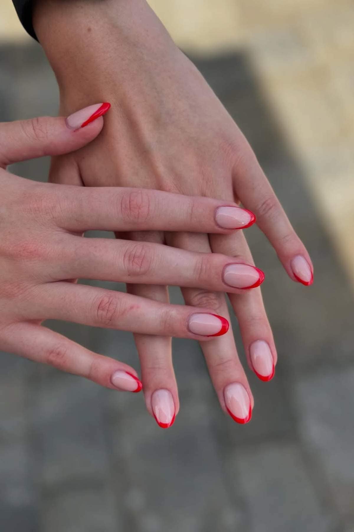 Two hands with almond-shaped nails featuring a natural base color and bright red tips, shown against a blurred outdoor background.