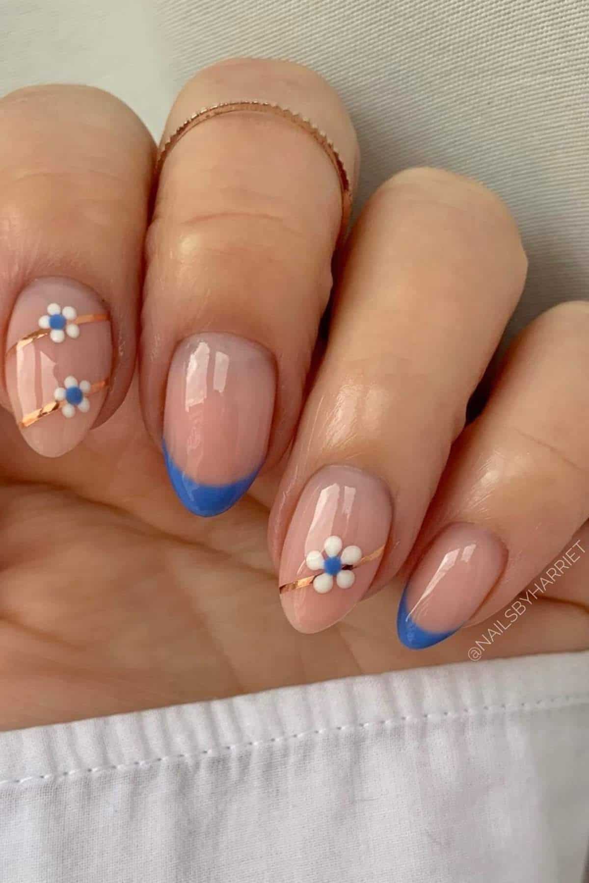 A hand with a nude manicure featuring blue French tips and white daisy flower designs with gold lines on two nails.