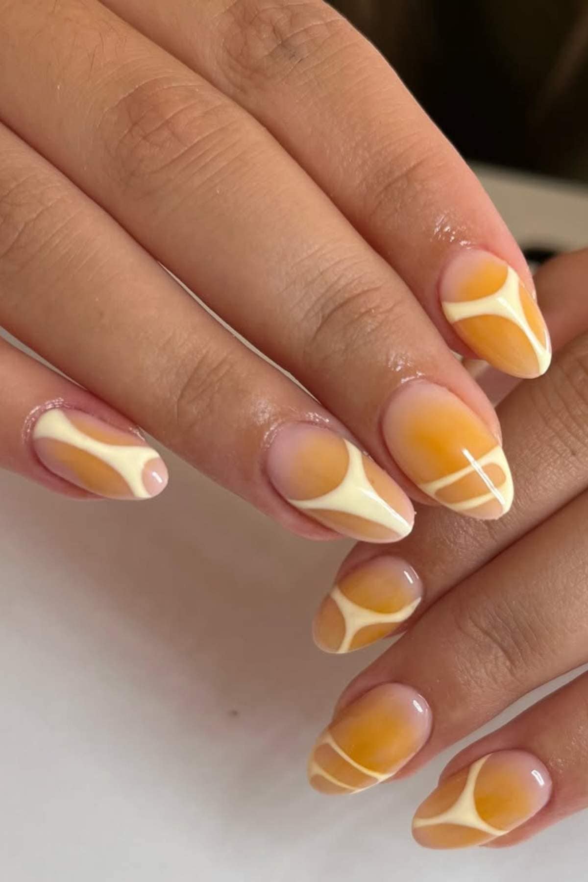 Close-up of hands with almond-shaped nails painted in yellow and white abstract geometric patterns on a neutral background.