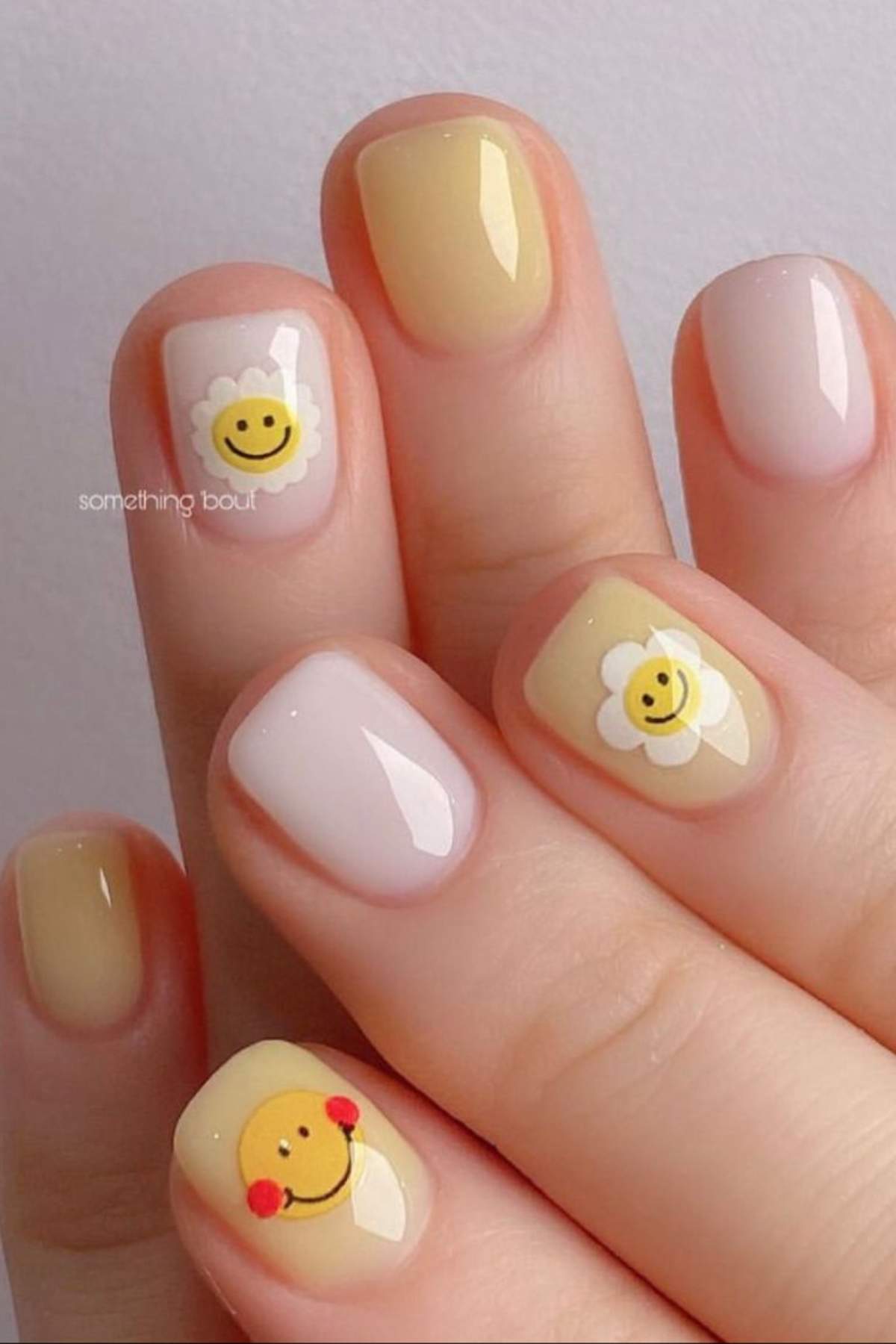 Close-up of hands with manicured nails in pastel yellow and nude shades, featuring smiley face and flower designs on some nails.