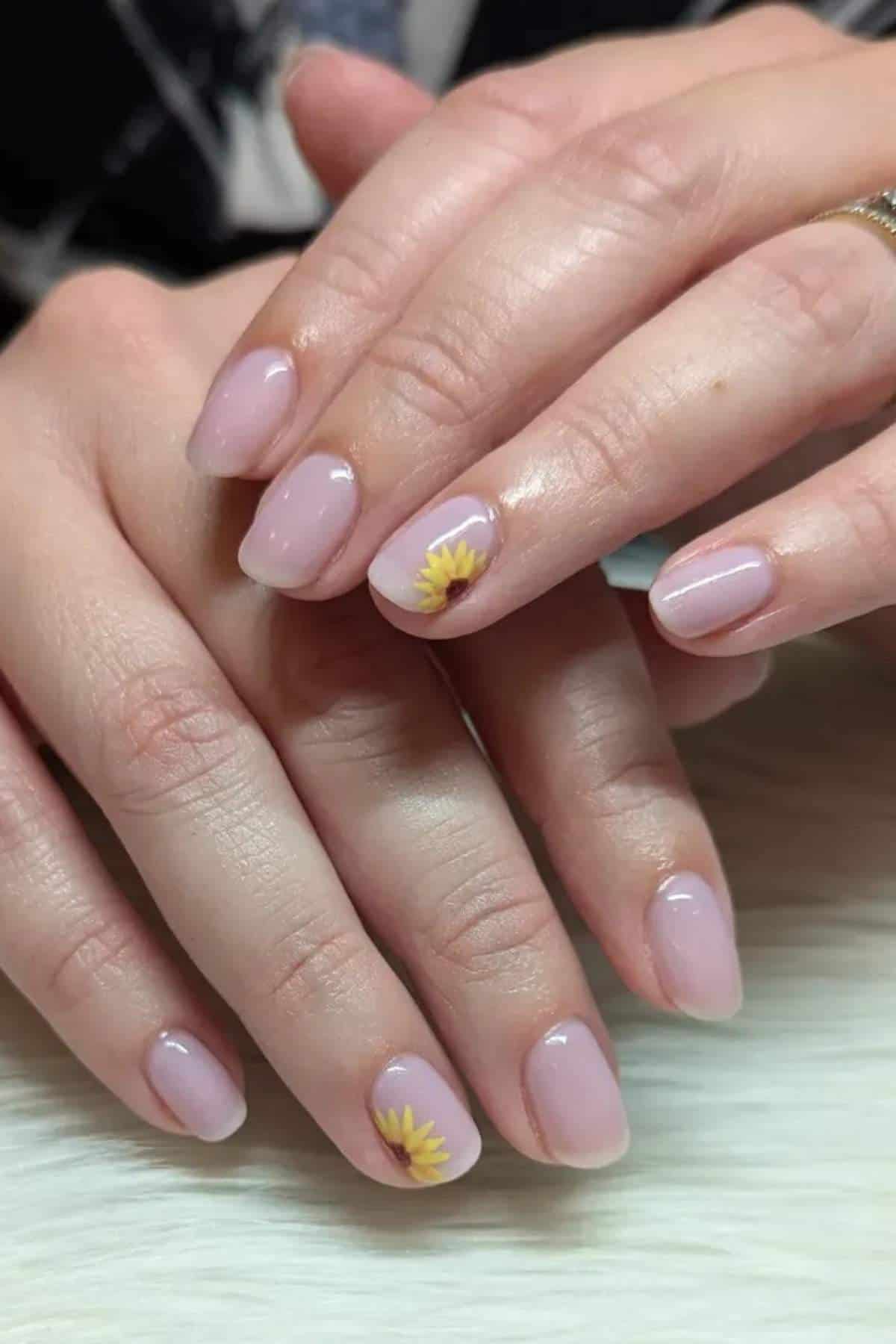Hands with short, oval-shaped nails painted light pink; ring finger nail on each hand features a small yellow flower design near the cuticle.