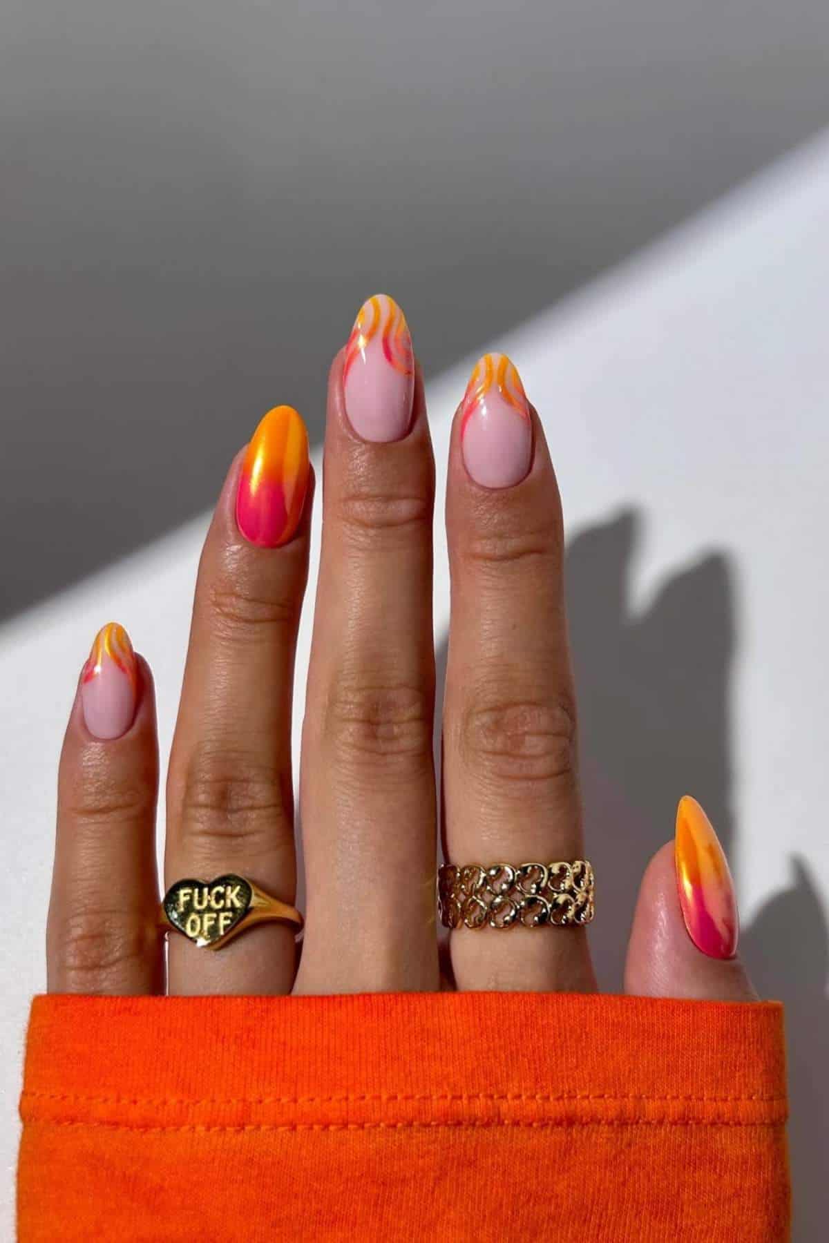 A hand with long, almond-shaped nails painted in pink, orange, and yellow swirls; the person wears two rings and an orange sleeve.