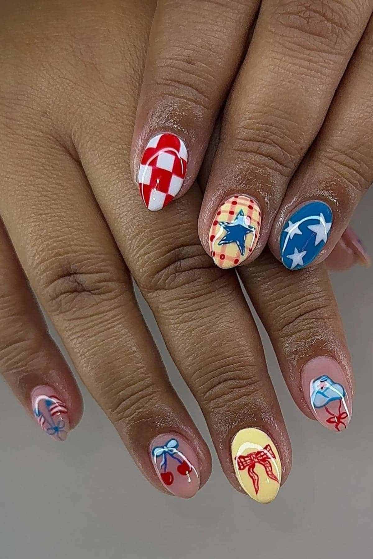 A hand with colorful nail art featuring various patterns, stars, bows, and checkerboard designs in red, blue, white, and yellow tones.