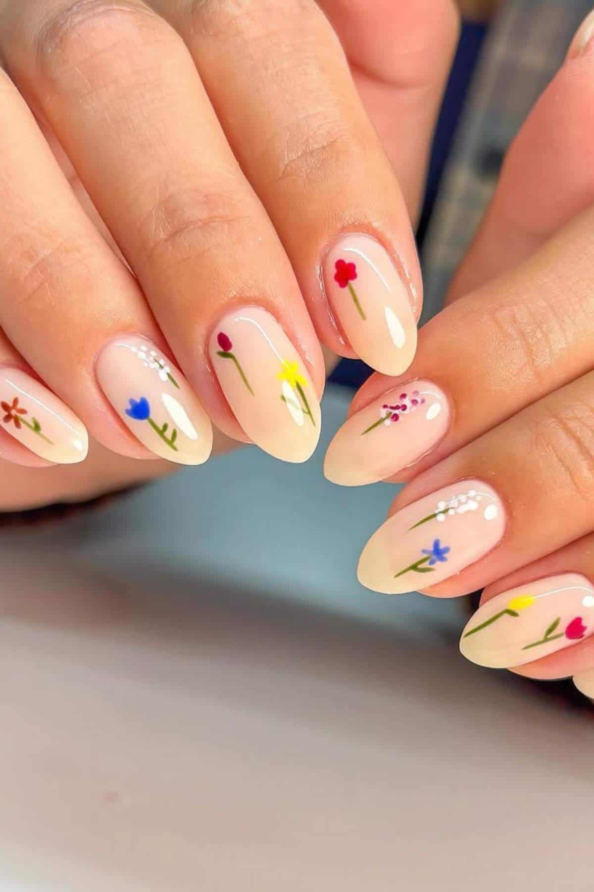 Close-up of hands with almond-shaped nails painted in a nude color, each decorated with a small, colorful flower design.