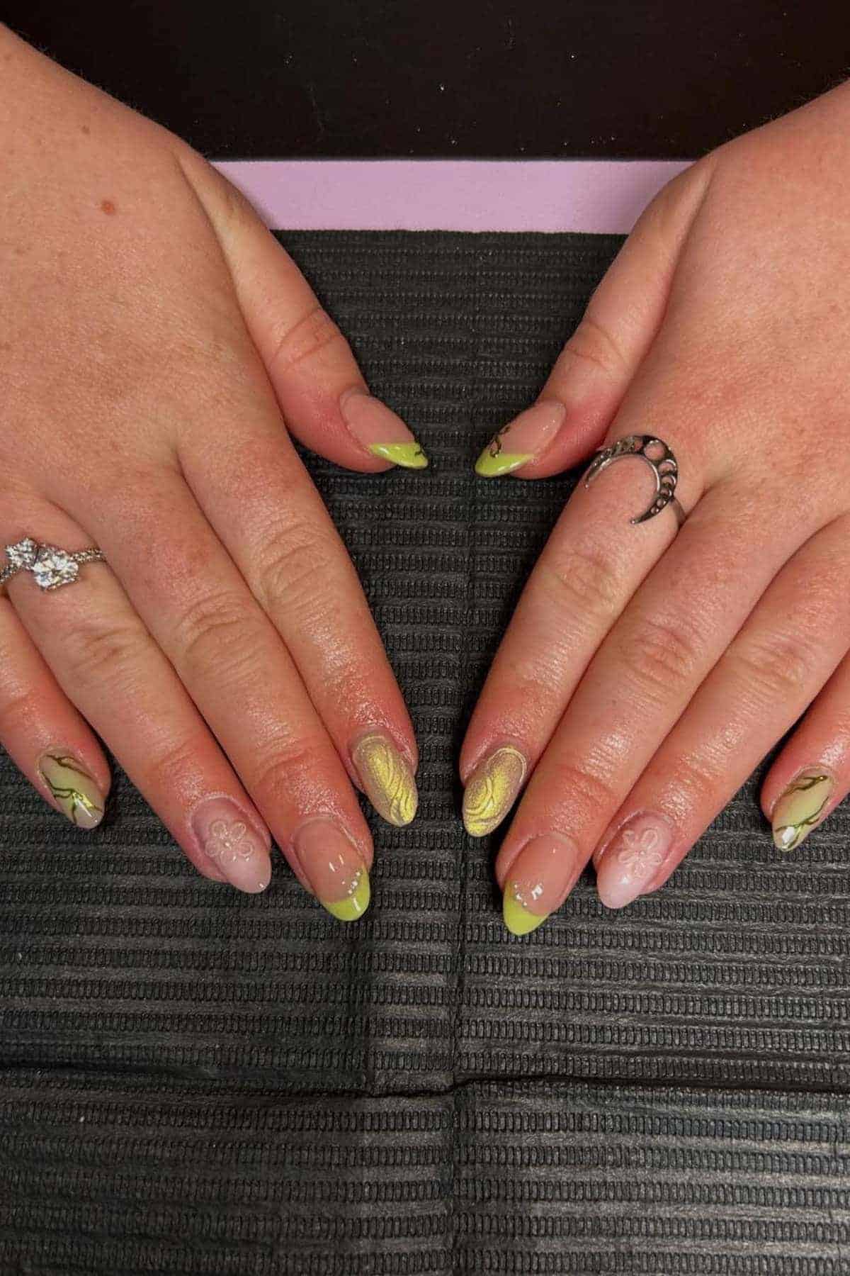 Two hands with almond-shaped nails featuring green and yellow abstract designs; rings are worn on both ring fingers.