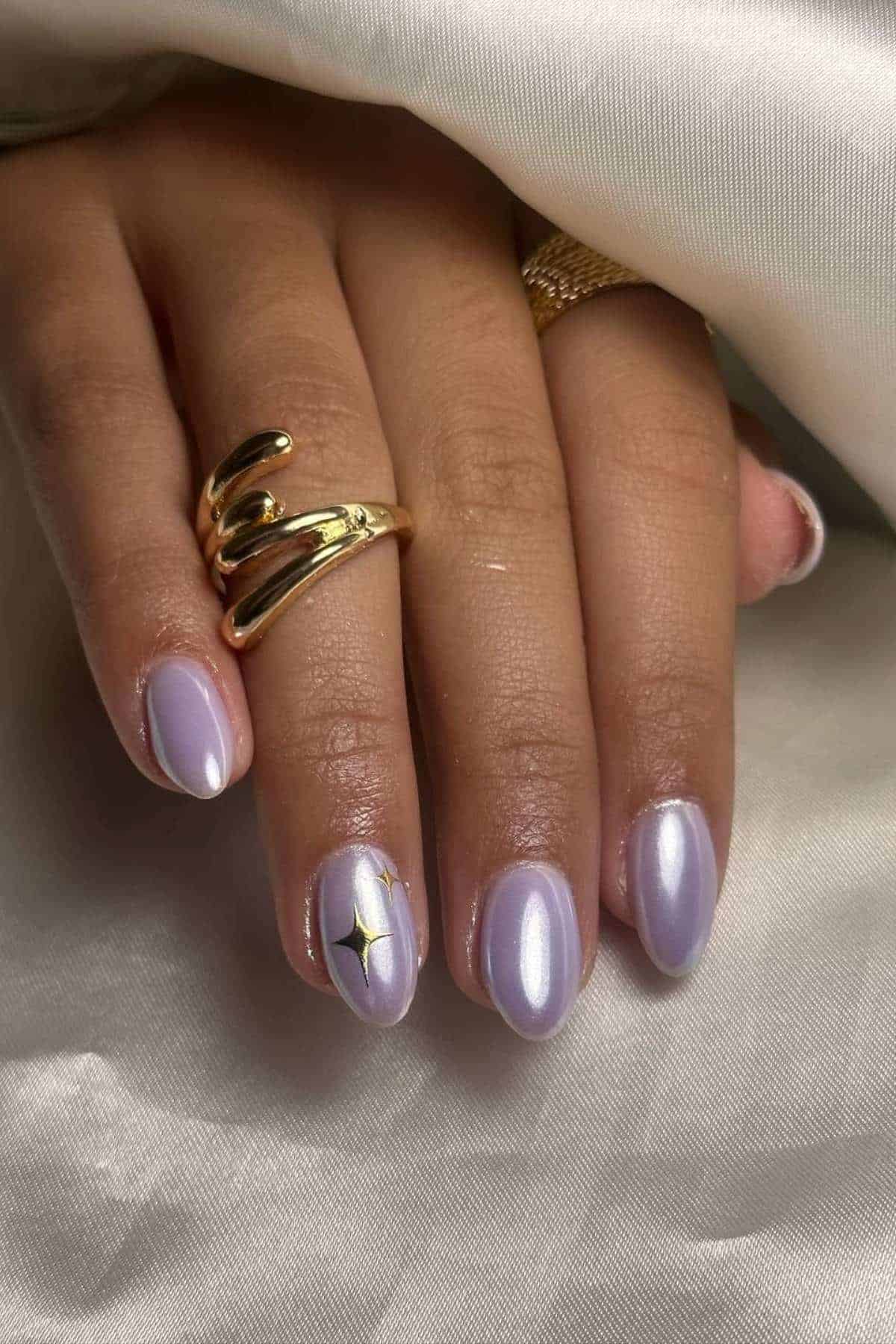 A hand with light purple, pearlescent manicured nails; the ring finger features a small black and gold star design. The person is wearing two gold rings.