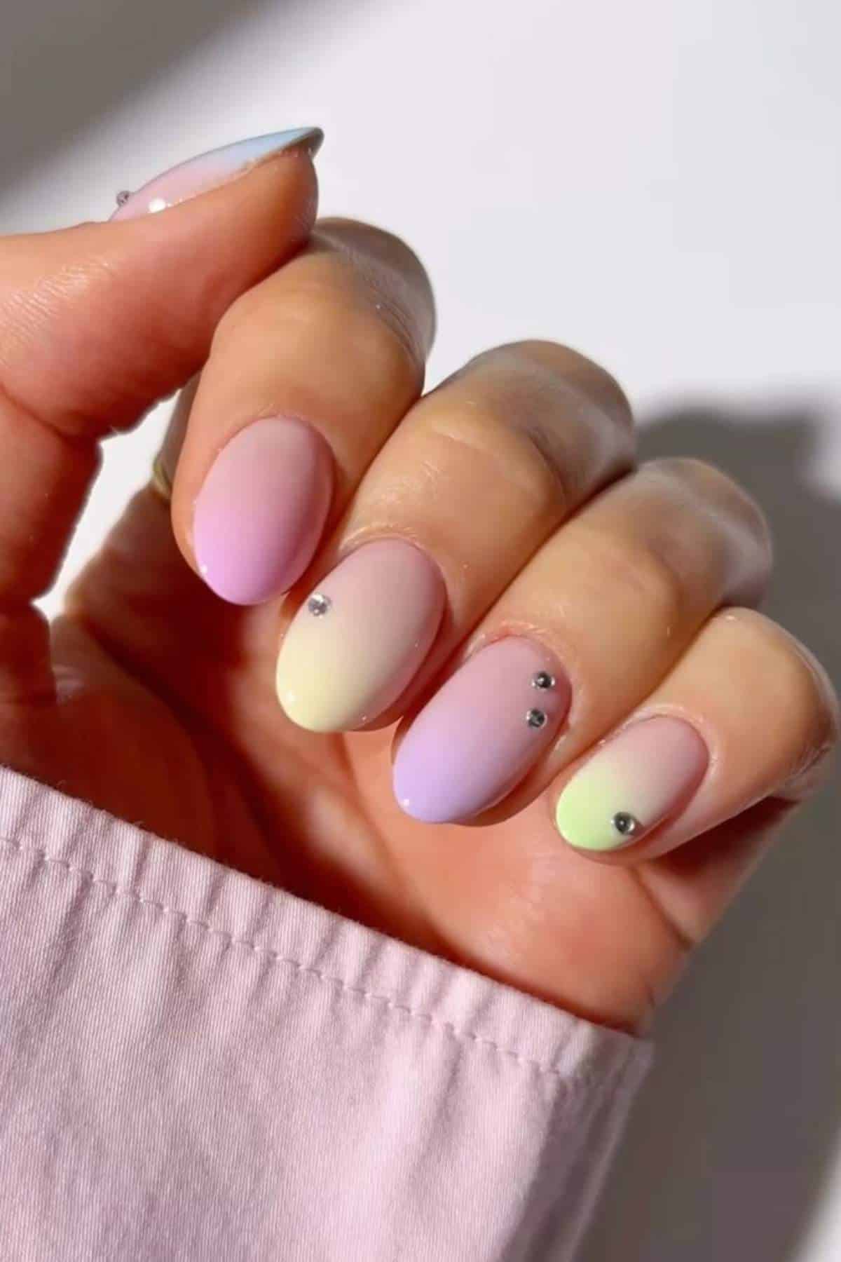 A hand with oval-shaped nails painted in pastel gradient colors and small rhinestone accents, against a light background.