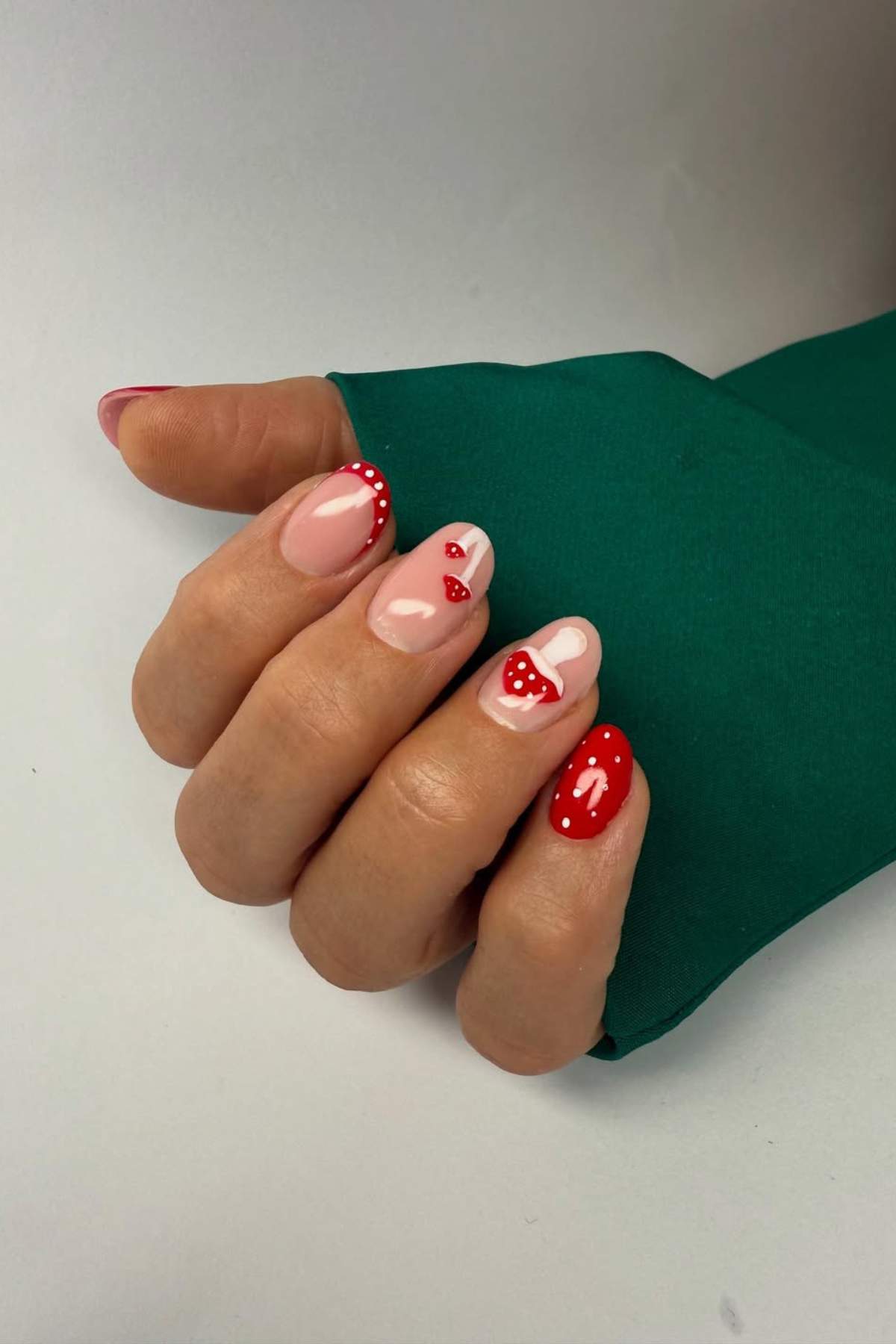 A hand with manicured nails featuring pink and red polish, heart designs, and polka dots, resting on a green fabric background.
